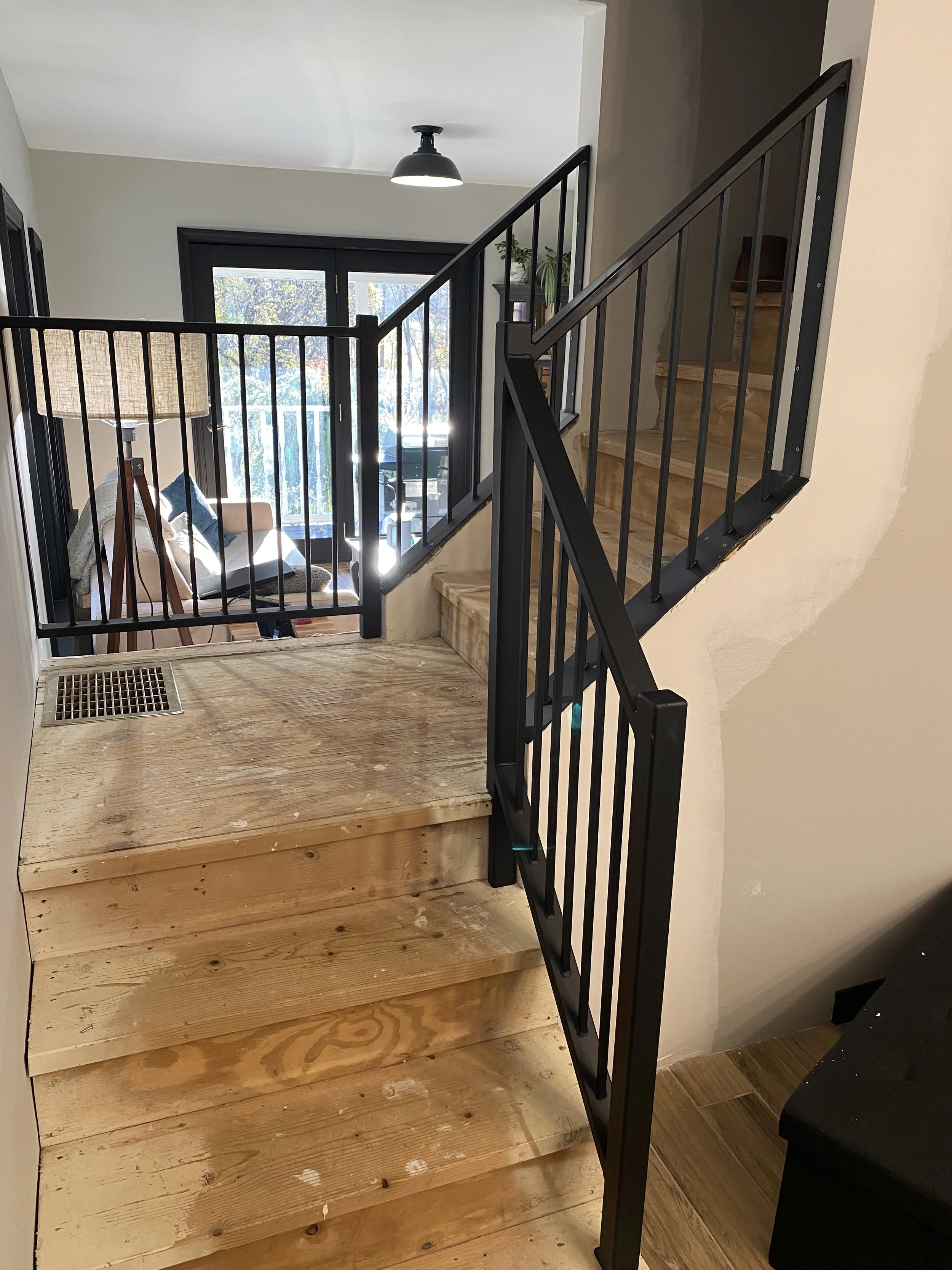 Interior view of a staircase with unfinished wooden steps and black metal railing, leading to a sunlit living room with sliding glass doors and a sitting area.