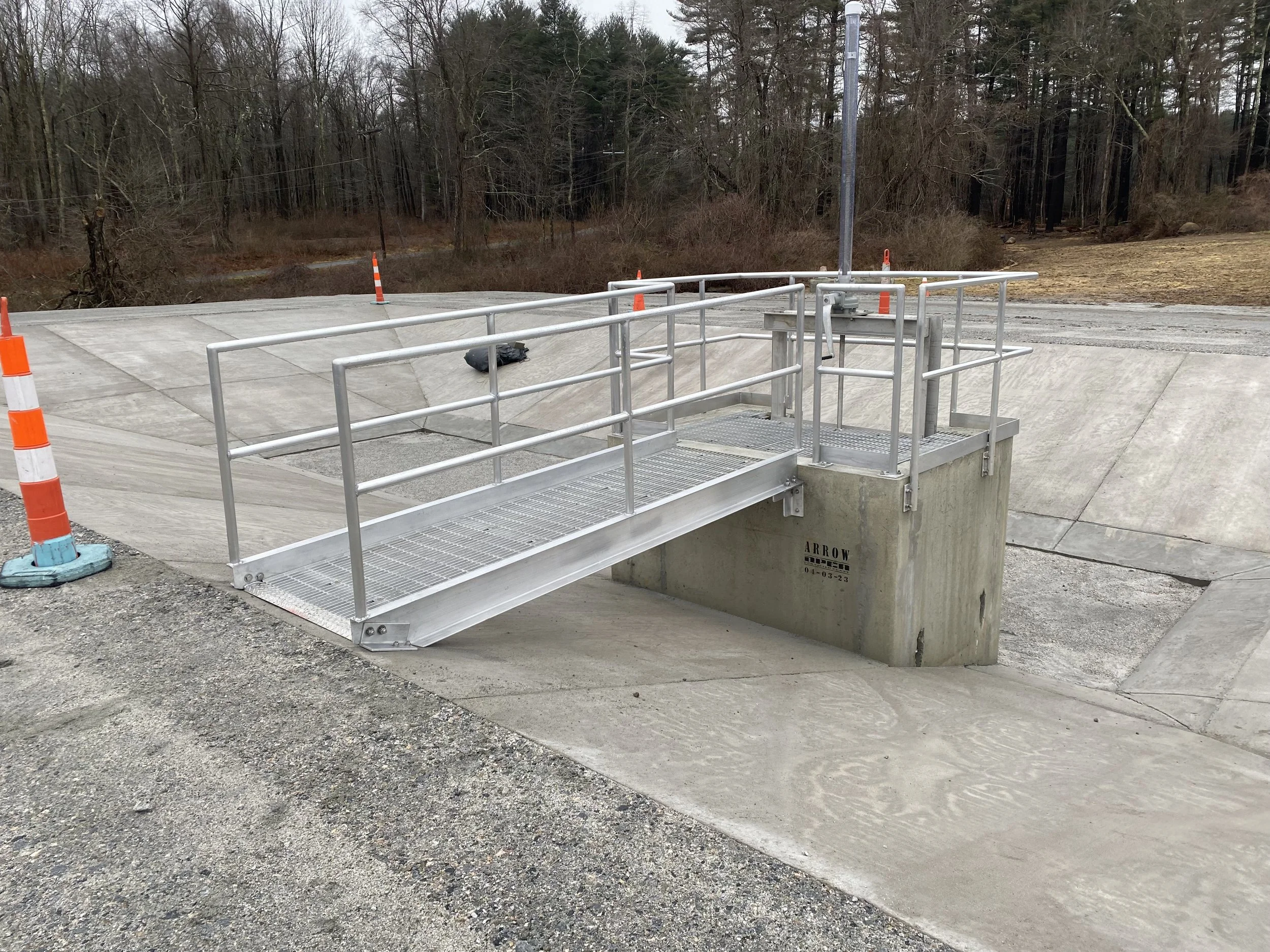 Metal access ramp with safety railing leading to a concrete structure at a drainage or water management site, with orange traffic cones in the background.