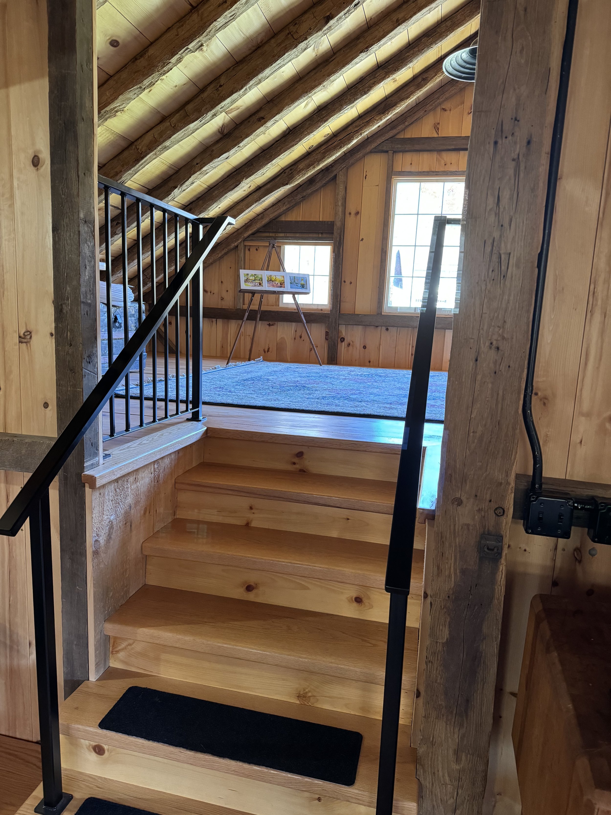 Wooden stairs leading up to a small loft space with two windows, wooden panel walls, and a blue rug, with a picture frame on an easel in the background.