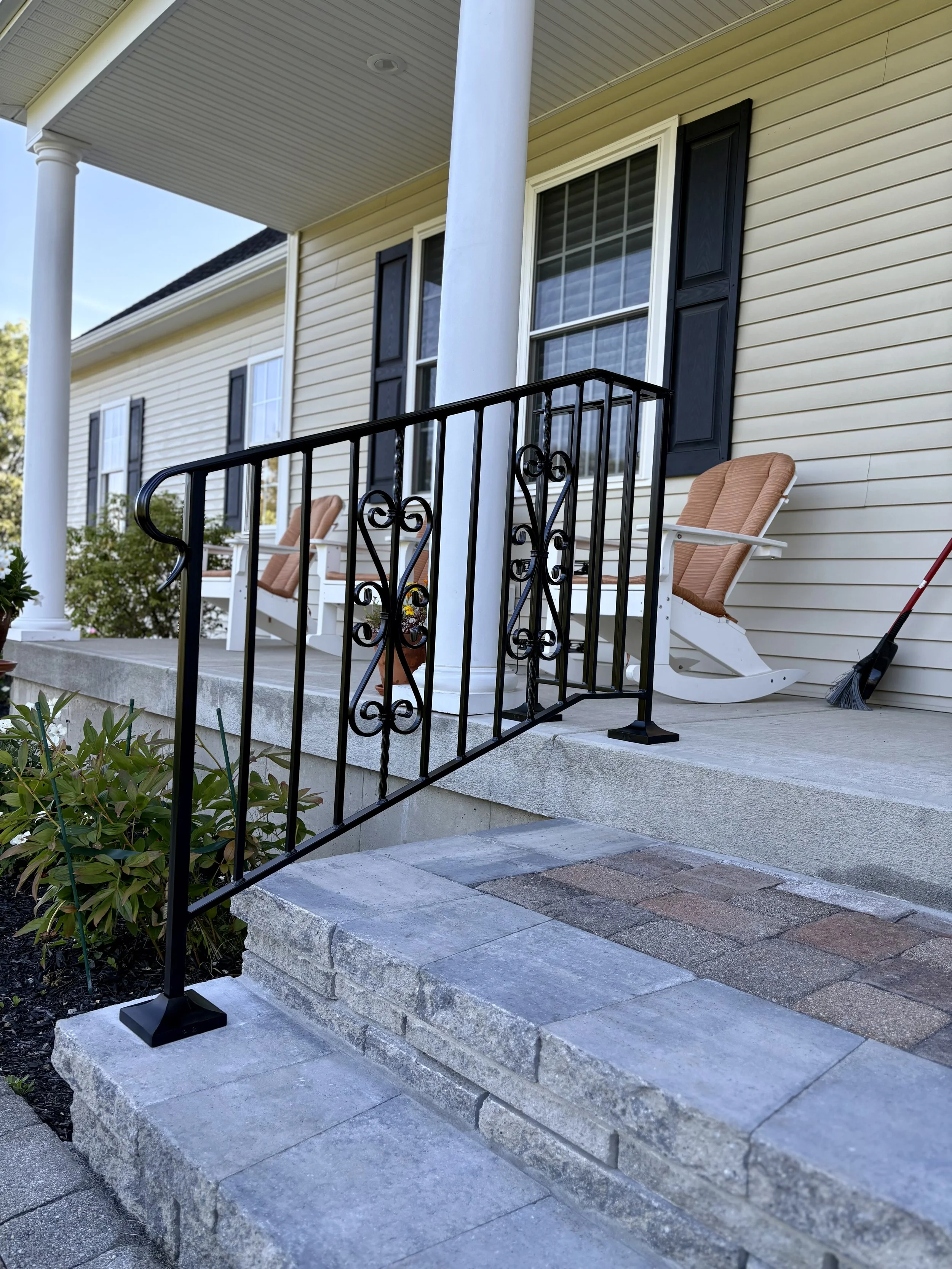 Front porch with steps, black wrought iron railing, outdoor chairs, a broom, and a house with beige siding and black shutters.