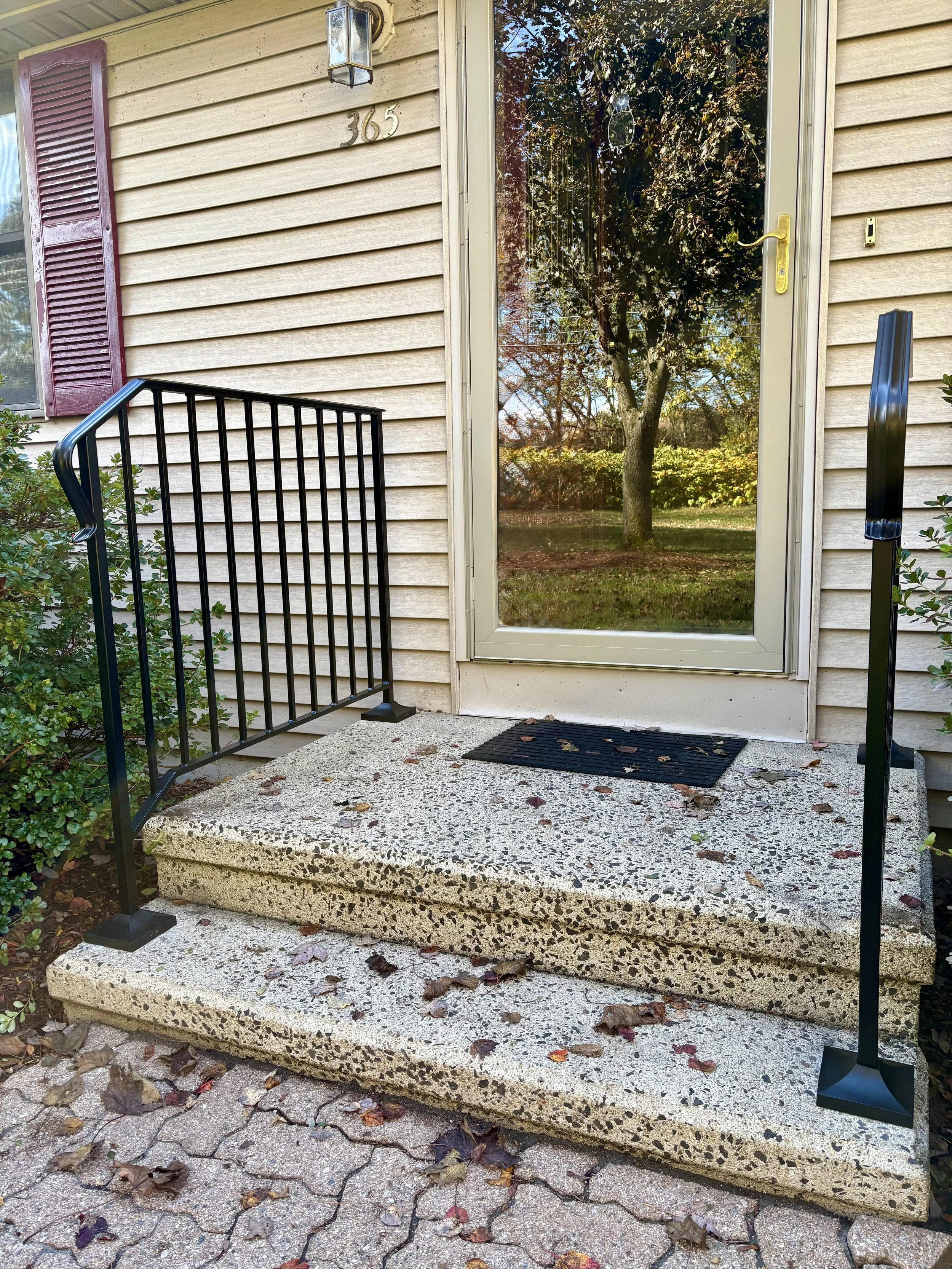 Concrete porch with two steps leading to a glass door with a yellow handle, black metal railings on either side, surrounded by fallen leaves, outside a house with beige siding and purple shutters.