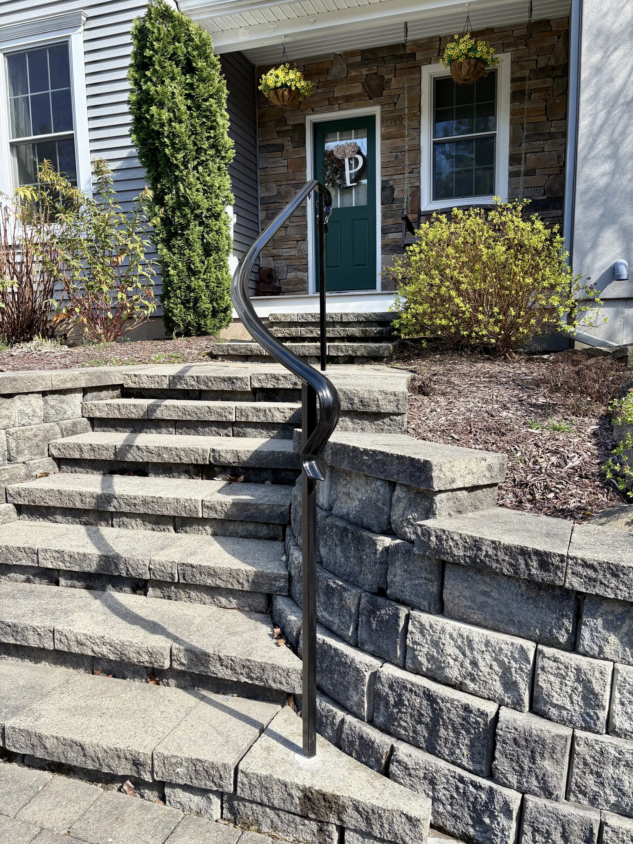 Stone steps leading to a front porch with a green door, surrounded by bushes, hanging flower baskets, and a tall tree.