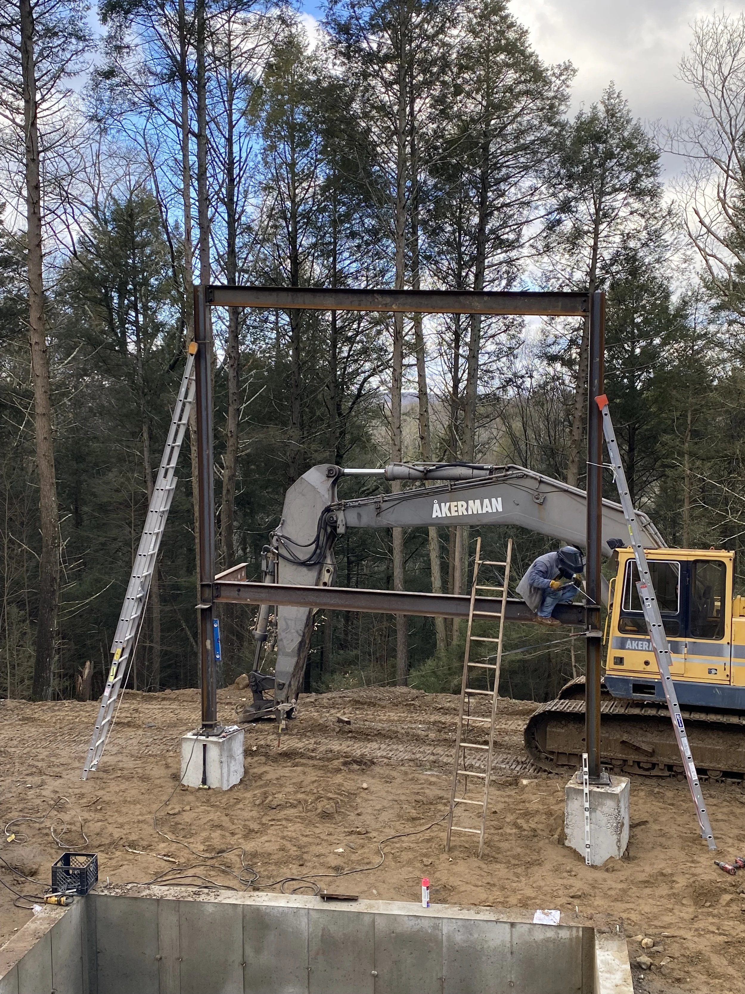 Construction site with a worker on an excavator, with ladders and steel beams being assembled in a forested area.