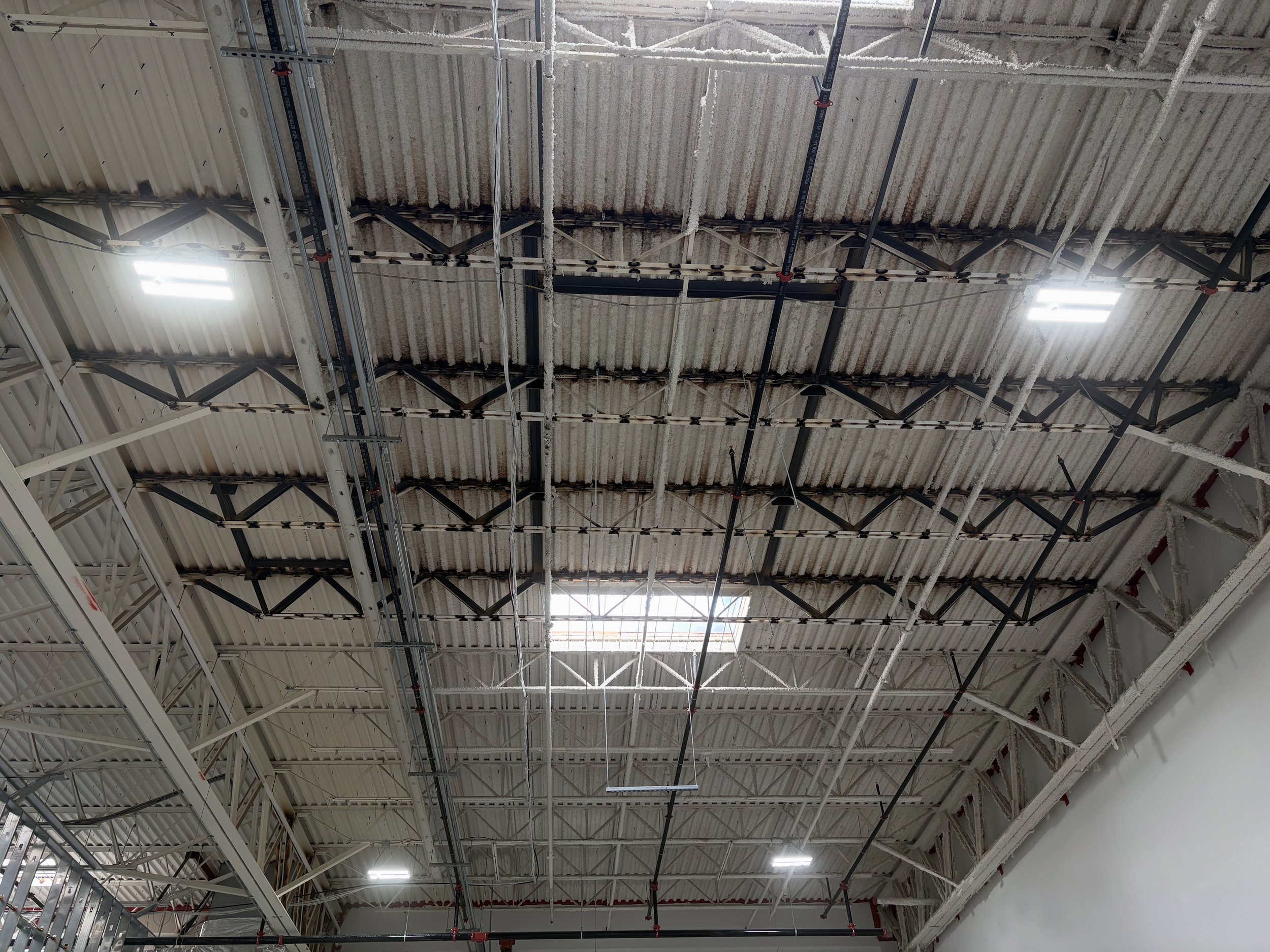 Interior view of a large industrial or commercial building ceiling, showing exposed metal framework, pipes, and insulation. Fluorescent lights are mounted on the ceiling.