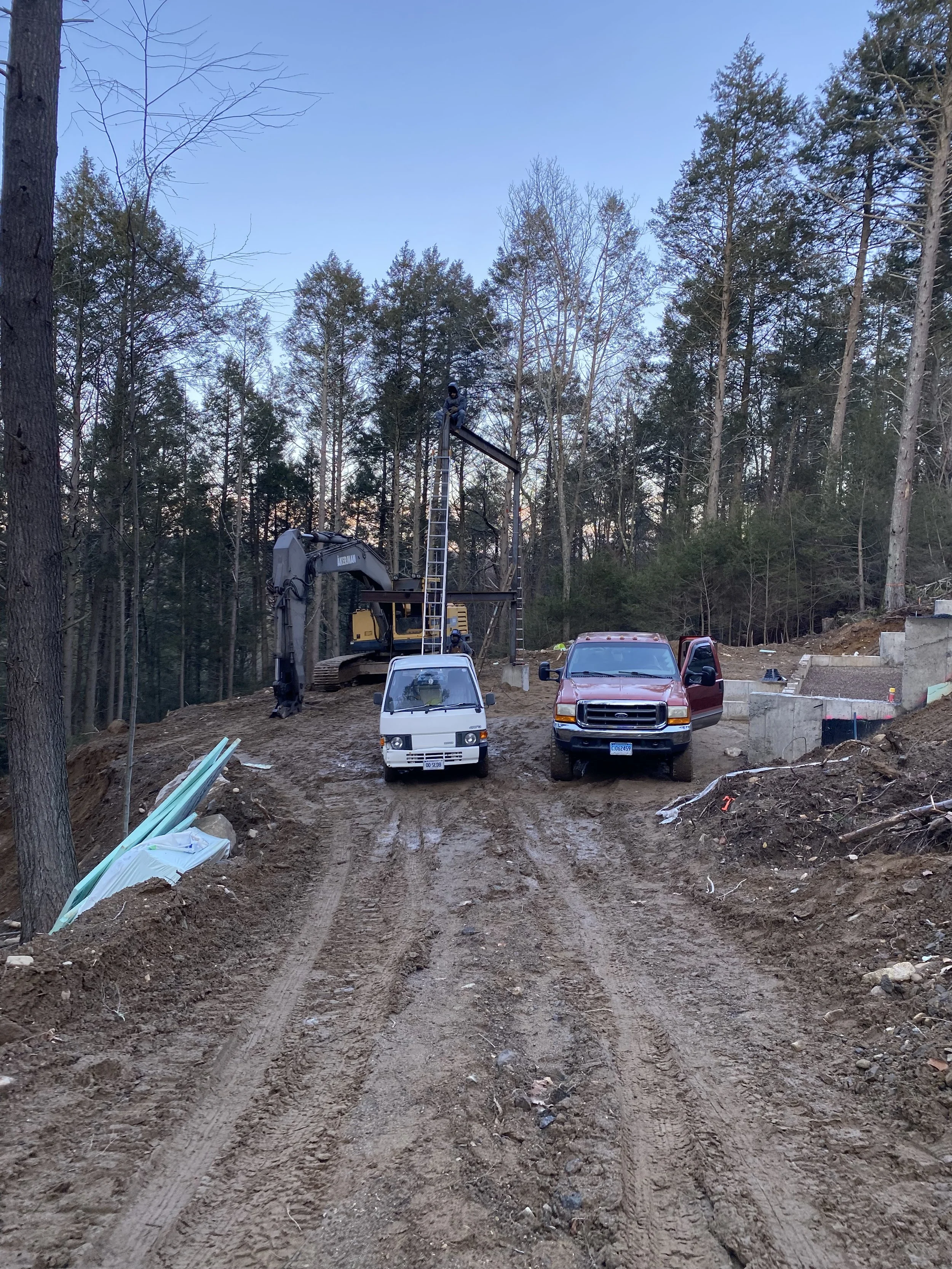 Construction site with trucks and heavy machinery on a dirt path surrounded by trees.