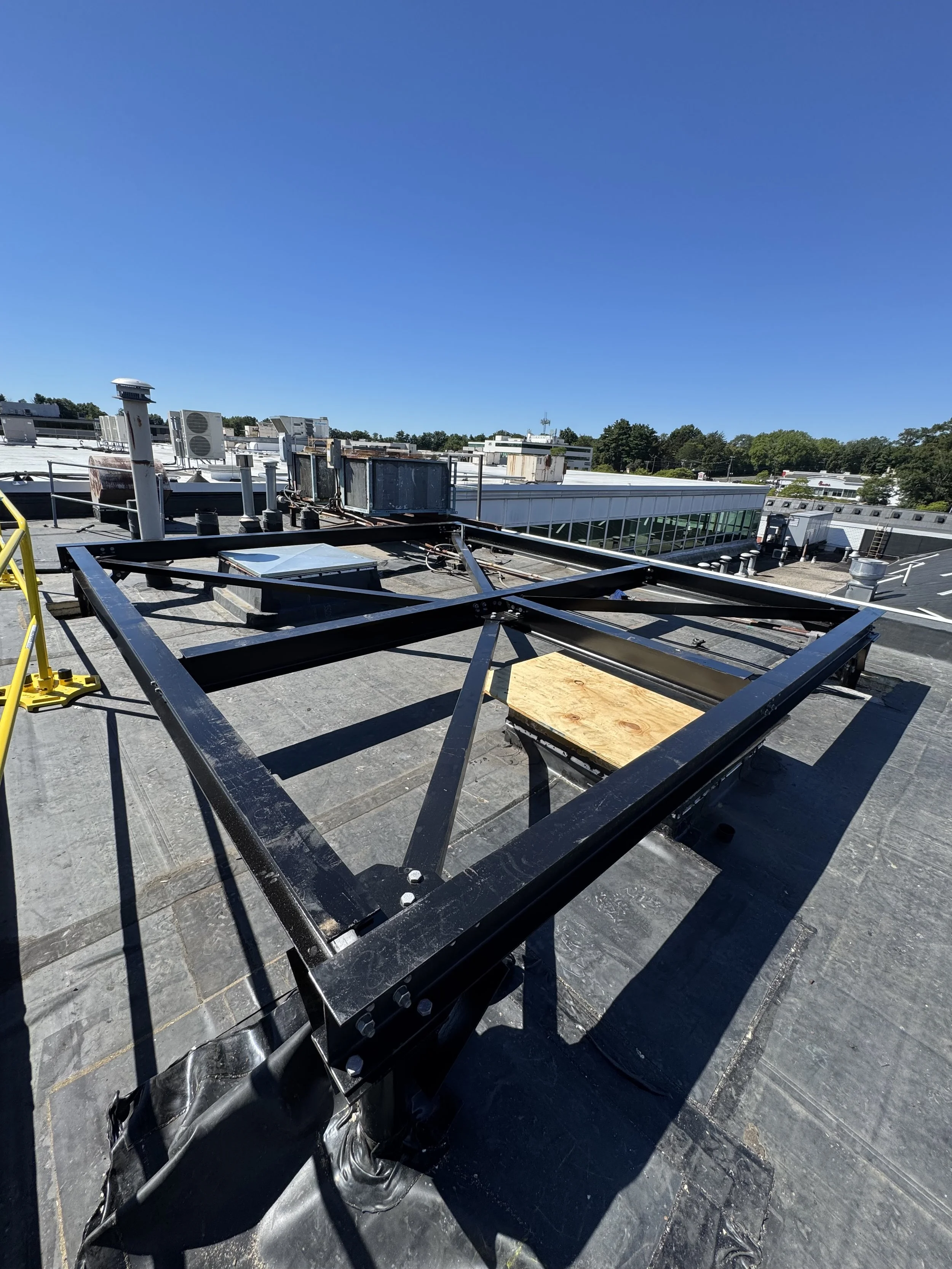 View from a rooftop showing a black metal frame structure under construction, with some wooden panels, HVAC units, and vents visible, under a clear blue sky.