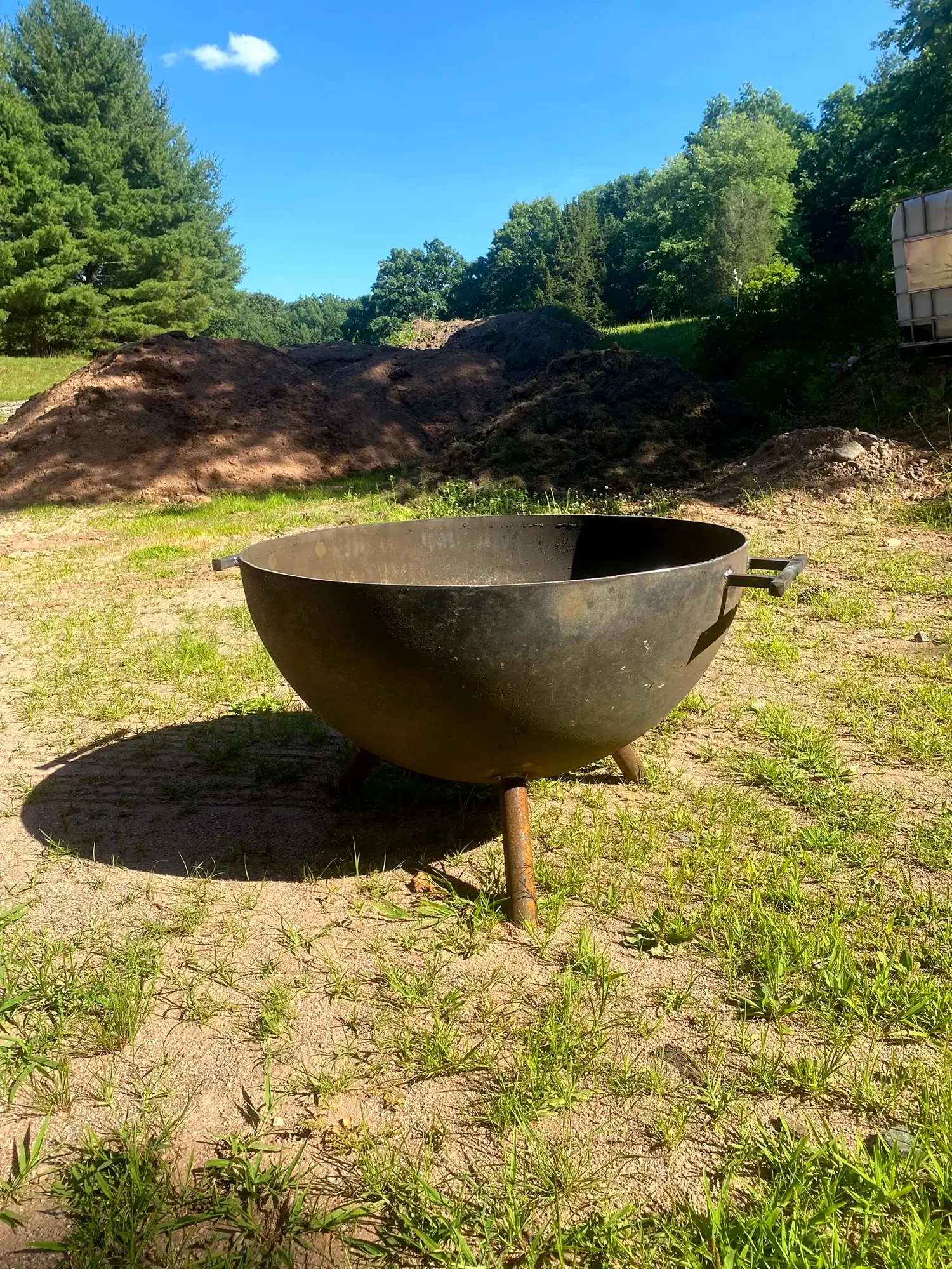 A black fire pit bowl on a grassy patch with a mound of dirt behind it, green trees, and a partly cloudy sky.