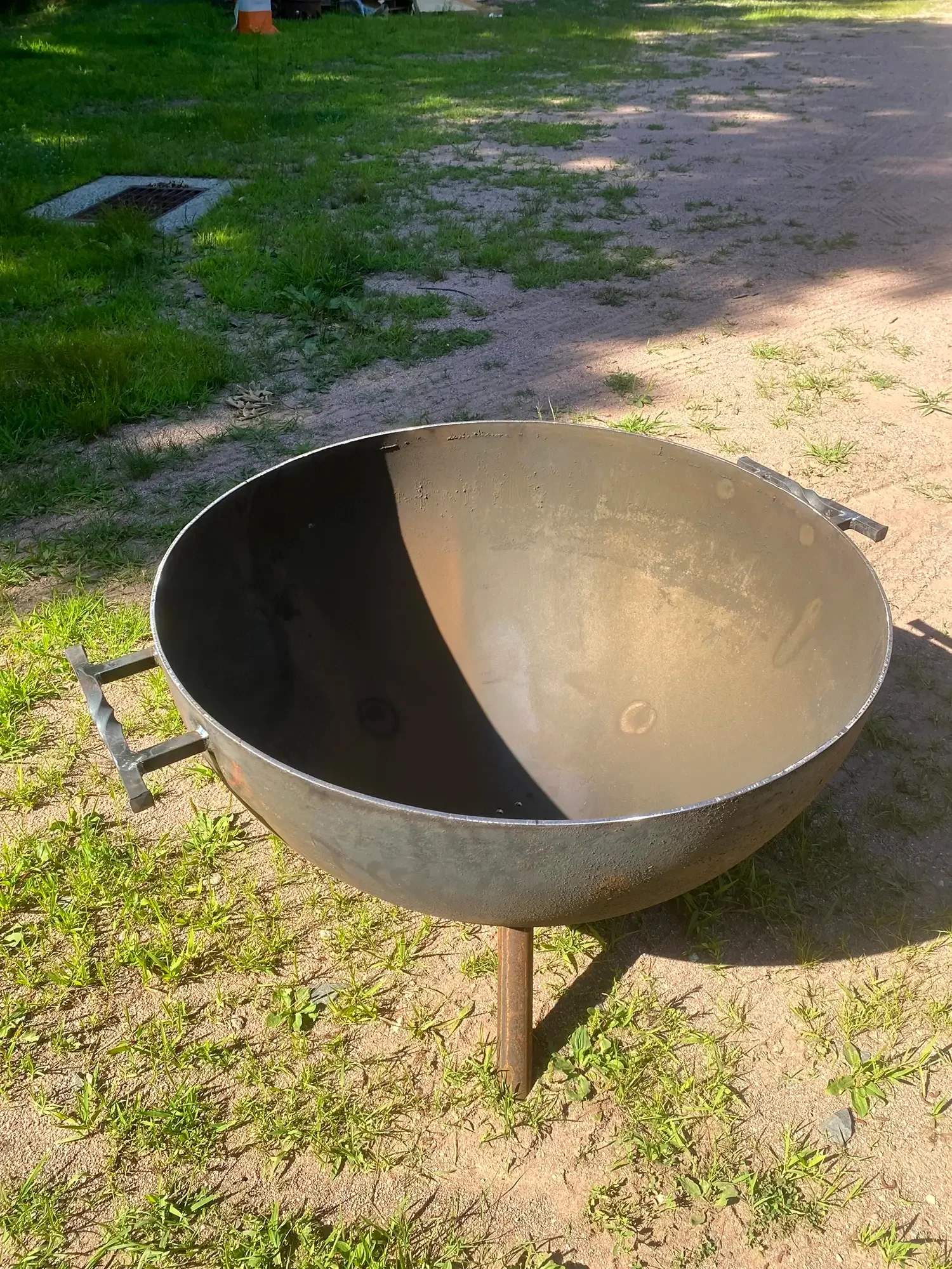 A round, rusted outdoor grill on a grassy and dirt area, with a wooden stake supporting it. In the background, there are patches of green grass, a drain, and some objects under a cover.