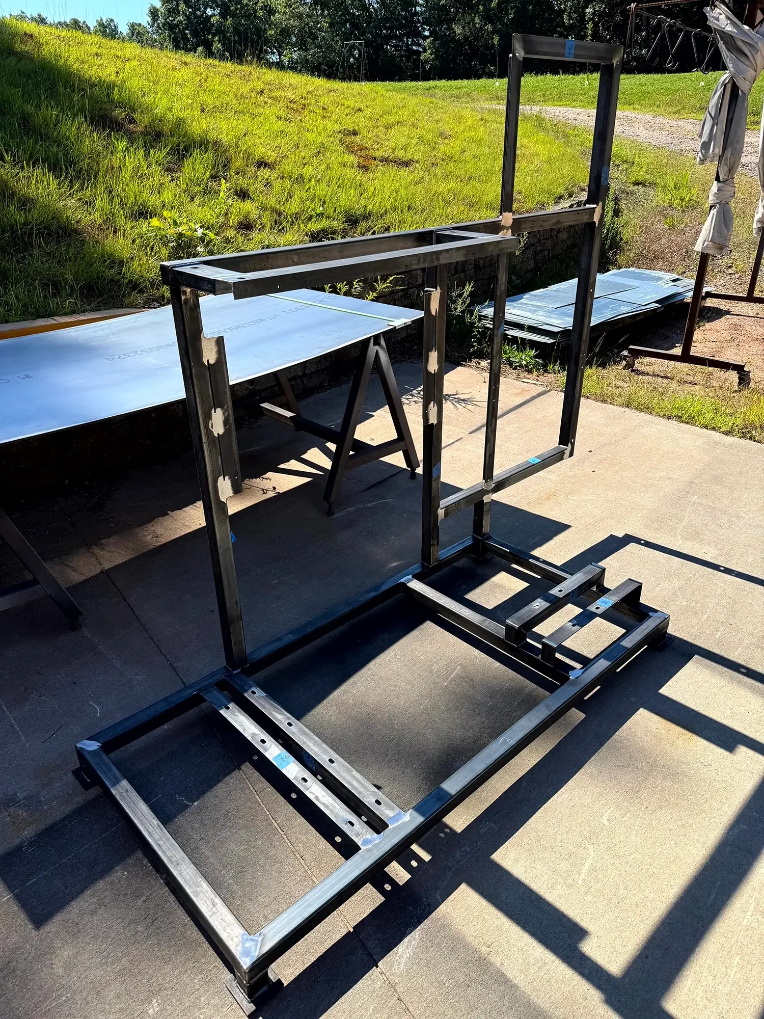 Metal frame structure being assembled outdoors, with a metal worktable and stacks of metal sheets in the background.
