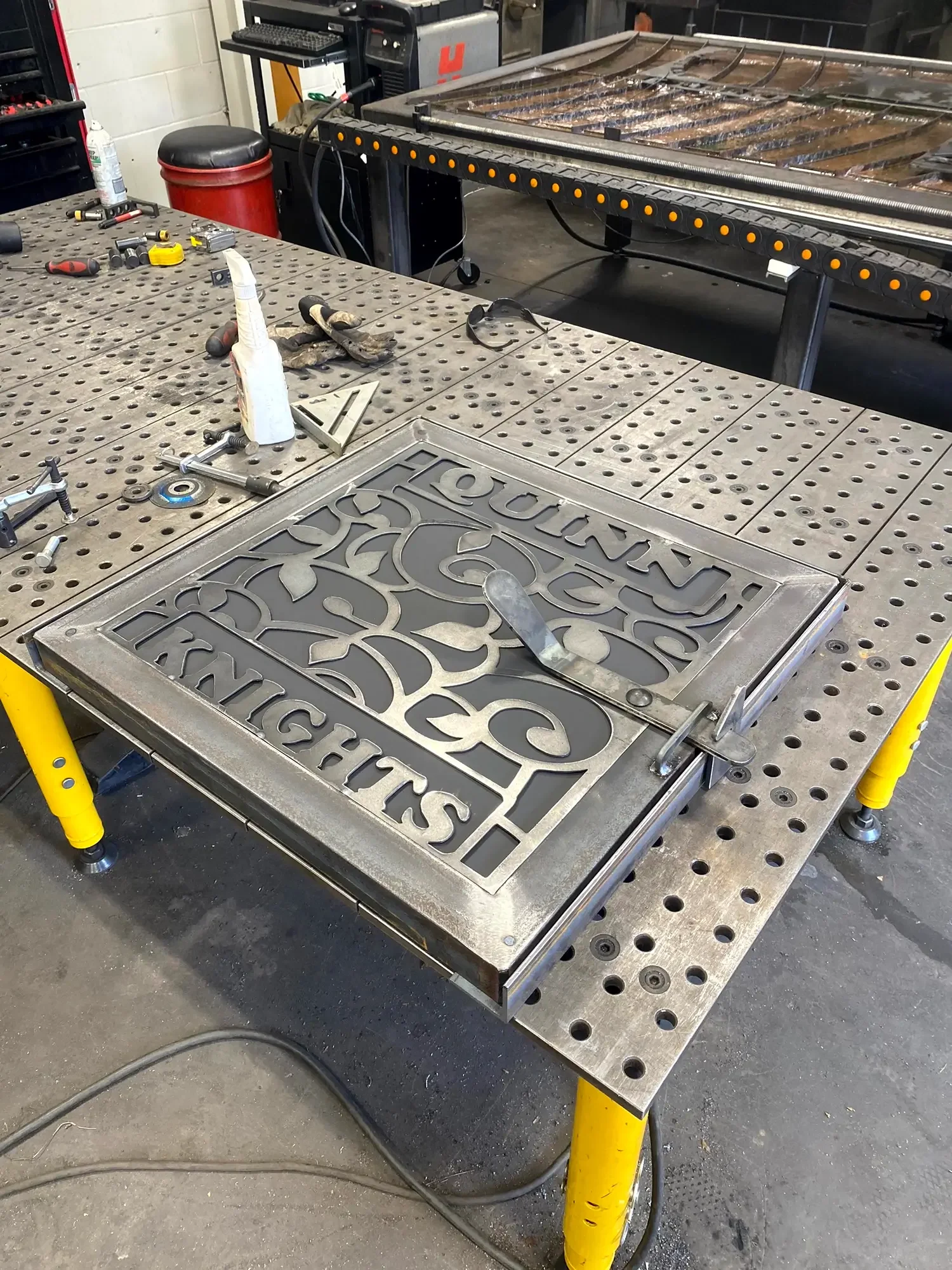 Workshop table with tools and gloves, metal ventilation cover with the words 'Housings International' cut out, in front of a large piece of machinery or equipment.