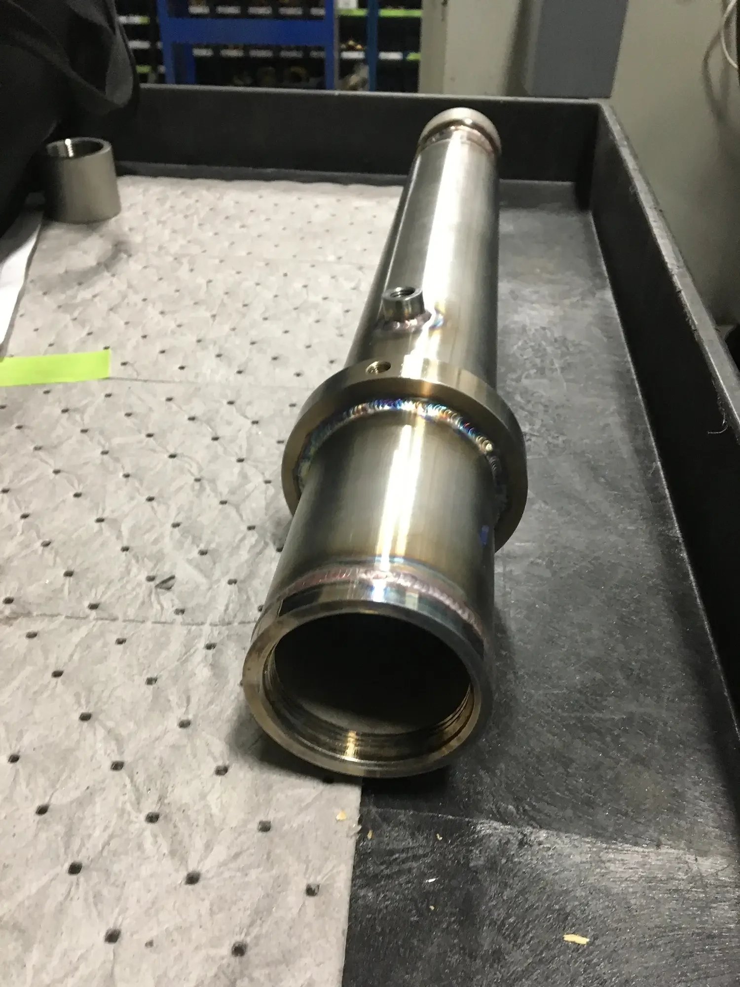 A cylindrical metal pipe or tube with a weld bead around one end, resting on a surface that is half gray rubber matting and half metal. In the background, there are shelves with items and a control panel or electrical box.