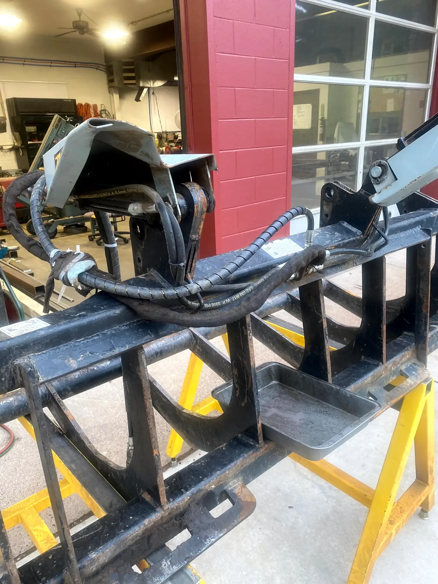 A metal frame for a machine or equipment, possibly a forklift or industrial lift, with various hydraulic hoses and components attached, placed on a yellow cart in an indoor workshop or industrial setting.