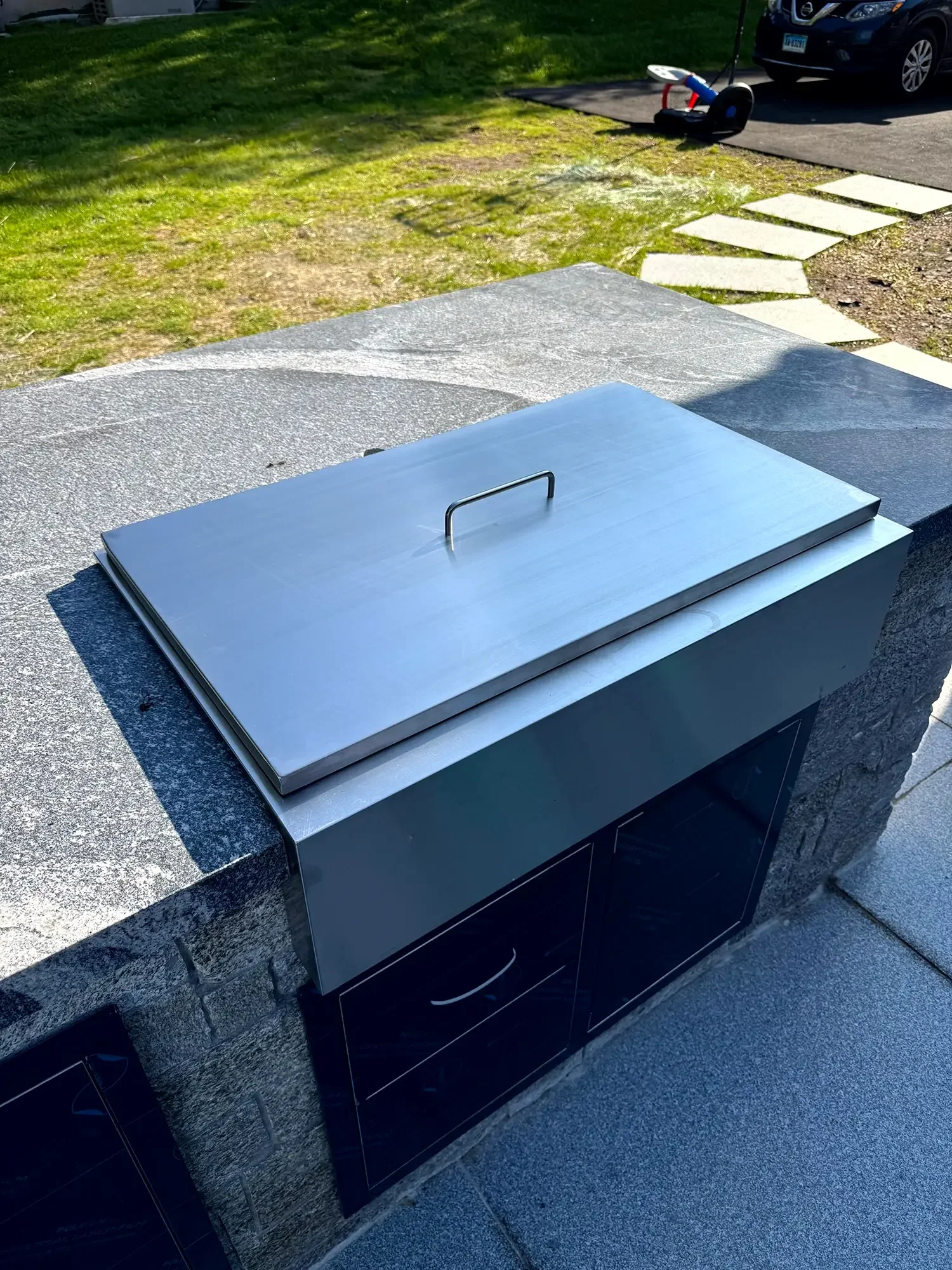 A stainless steel grill with a closed lid placed on a stone countertop outside. In the background, there is a grassy yard with a pathway of stepping stones, a lawnmower, and parked cars.