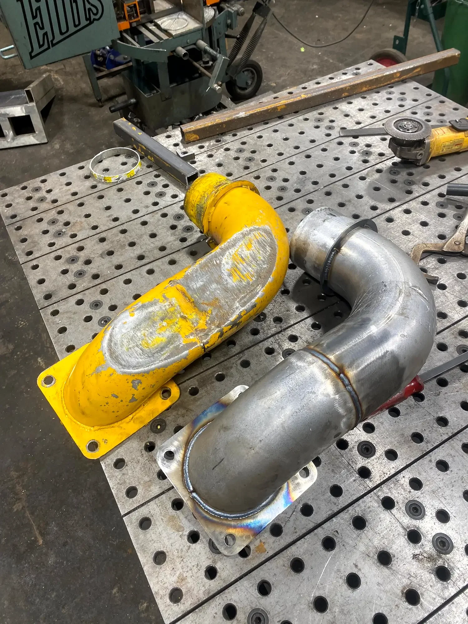 Metal pipes and fittings on a perforated metal workbench in a workshop setting, including a yellow pipe with welding marks and a silver pipe, surrounded by tools and equipment.