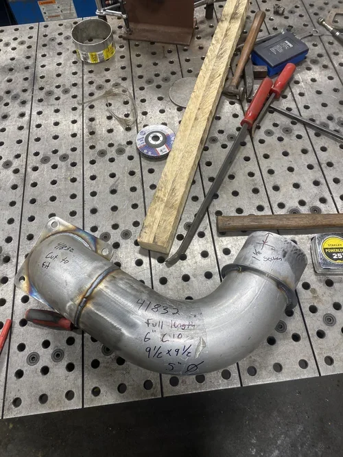 Metal pipe bent at a 90-degree angle on a perforated metal work table, with measurements and notes written on it, surrounded by tools and materials such as a ruler, a screwdriver, a wooden plank, and a metal ring.