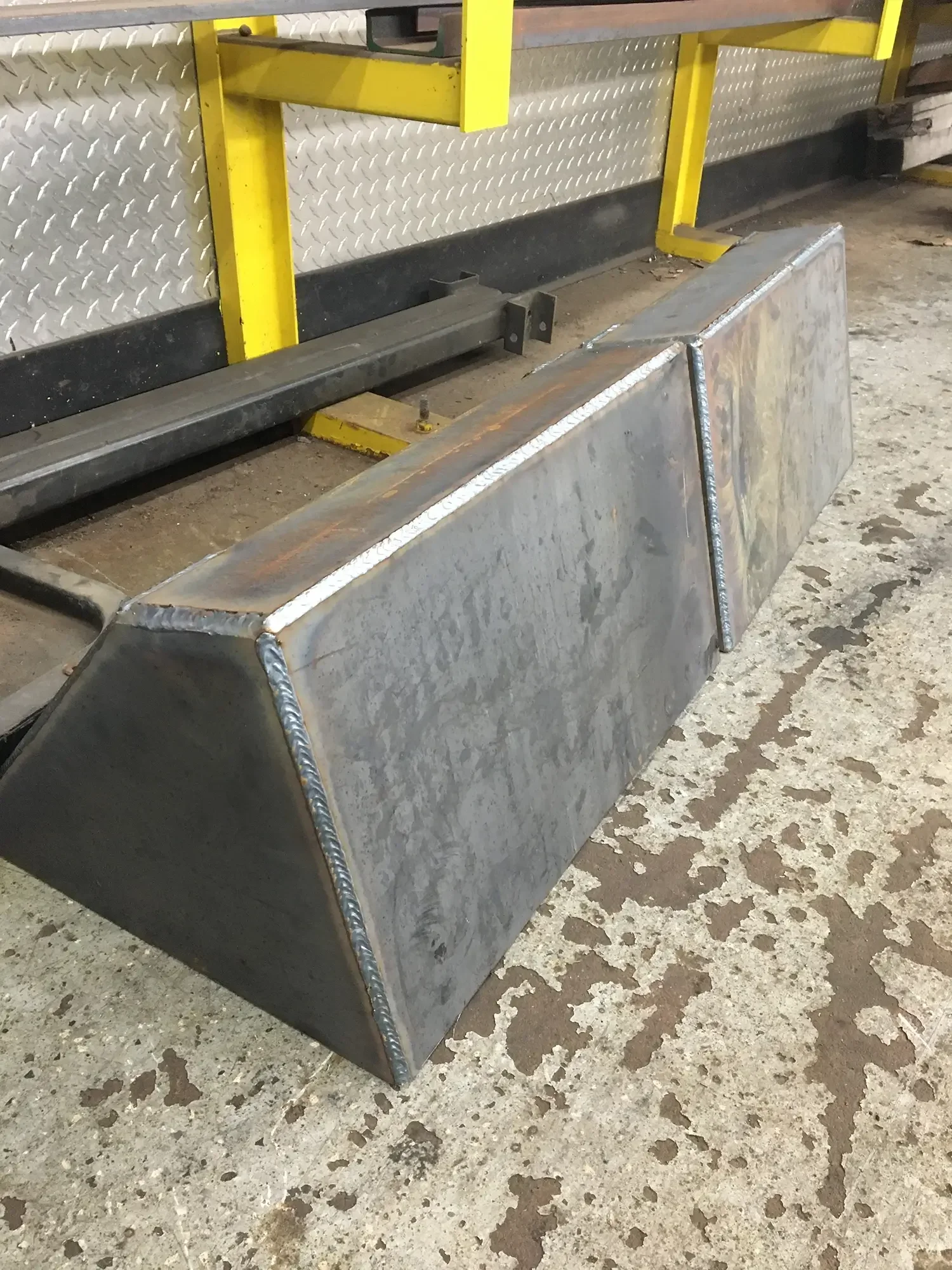 A large metal dumpster with welded seams, placed on a concrete floor with peeling paint. In the background, there is a metal wall with diamond plate pattern and yellow safety railing.