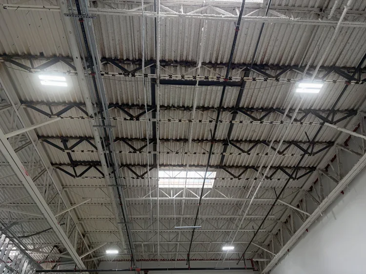 View of industrial warehouse ceiling with metal beams, tiles, lights, and some damage on the ceiling.