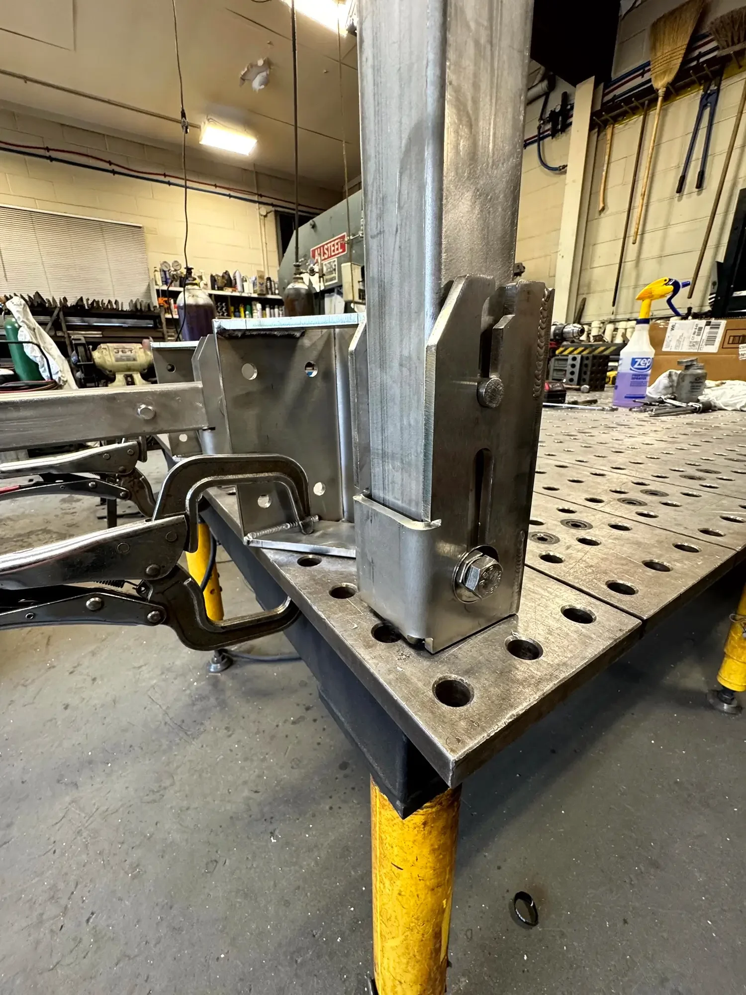 Close-up of a metal workbench with a metal jig or fixture attached, in a workshop with tools, spray bottle, and supplies visible in the background.