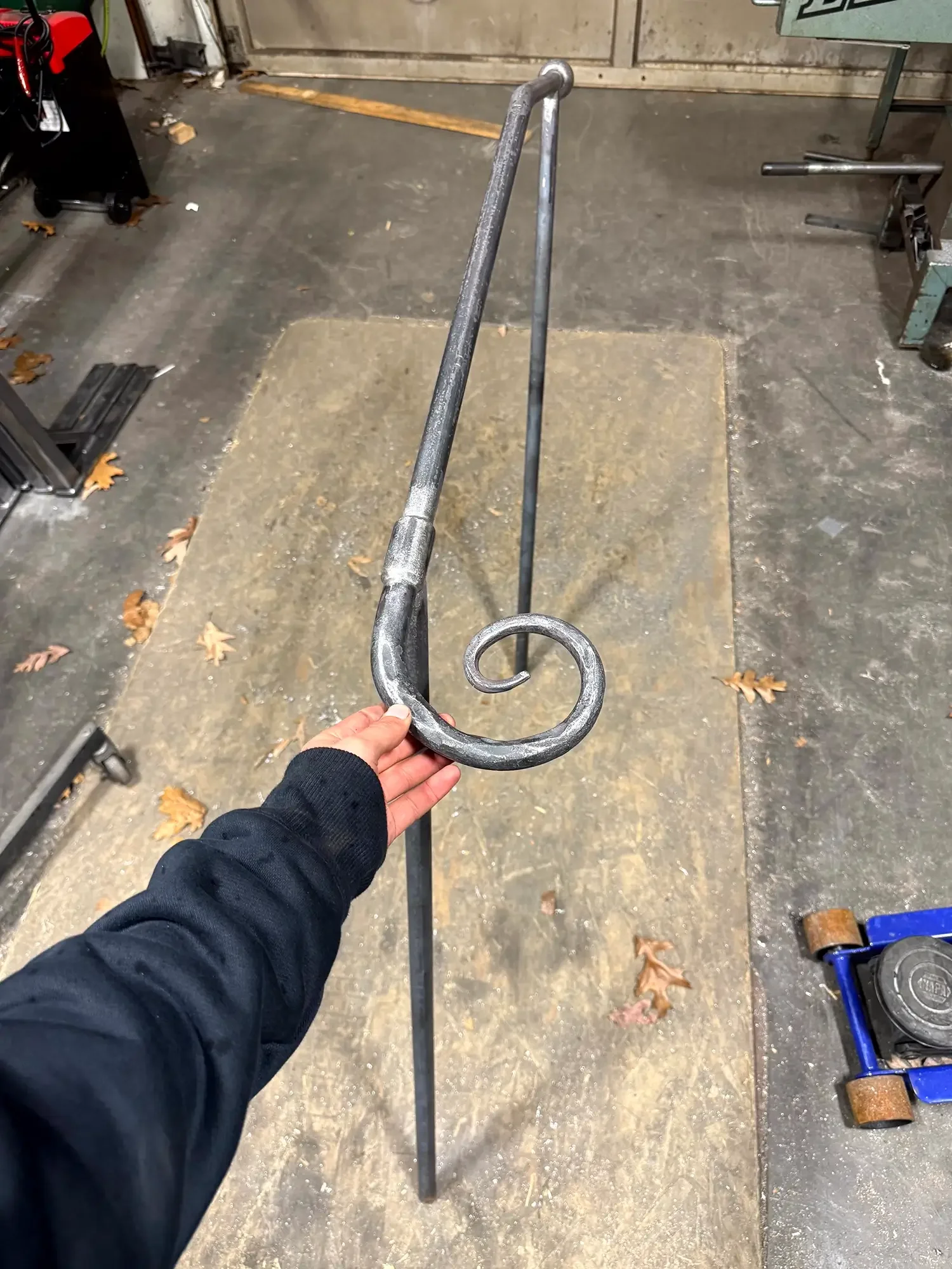 A person holding a metal decorative scroll with a hook at the end in a workshop or garage environment. The floor is concrete with scattered dry leaves. There are various tools and equipment around, including a wheelbarrow, a workbench, and items on t