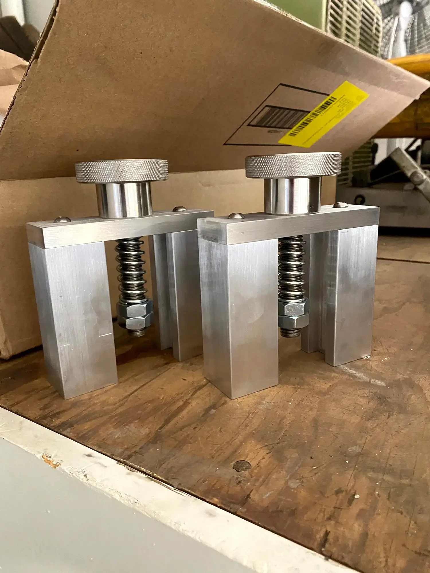 A metal mold with three cylindrical columns and twin helical springs on a wooden work surface, with a cardboard box in the background.