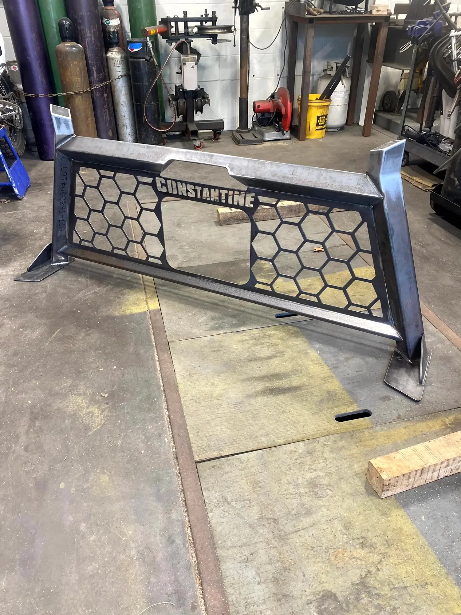 A metal car grille with the name 'Constantine' cut out, set on the floor inside a workshop or garage with tools, gas cylinders, and equipment in the background.