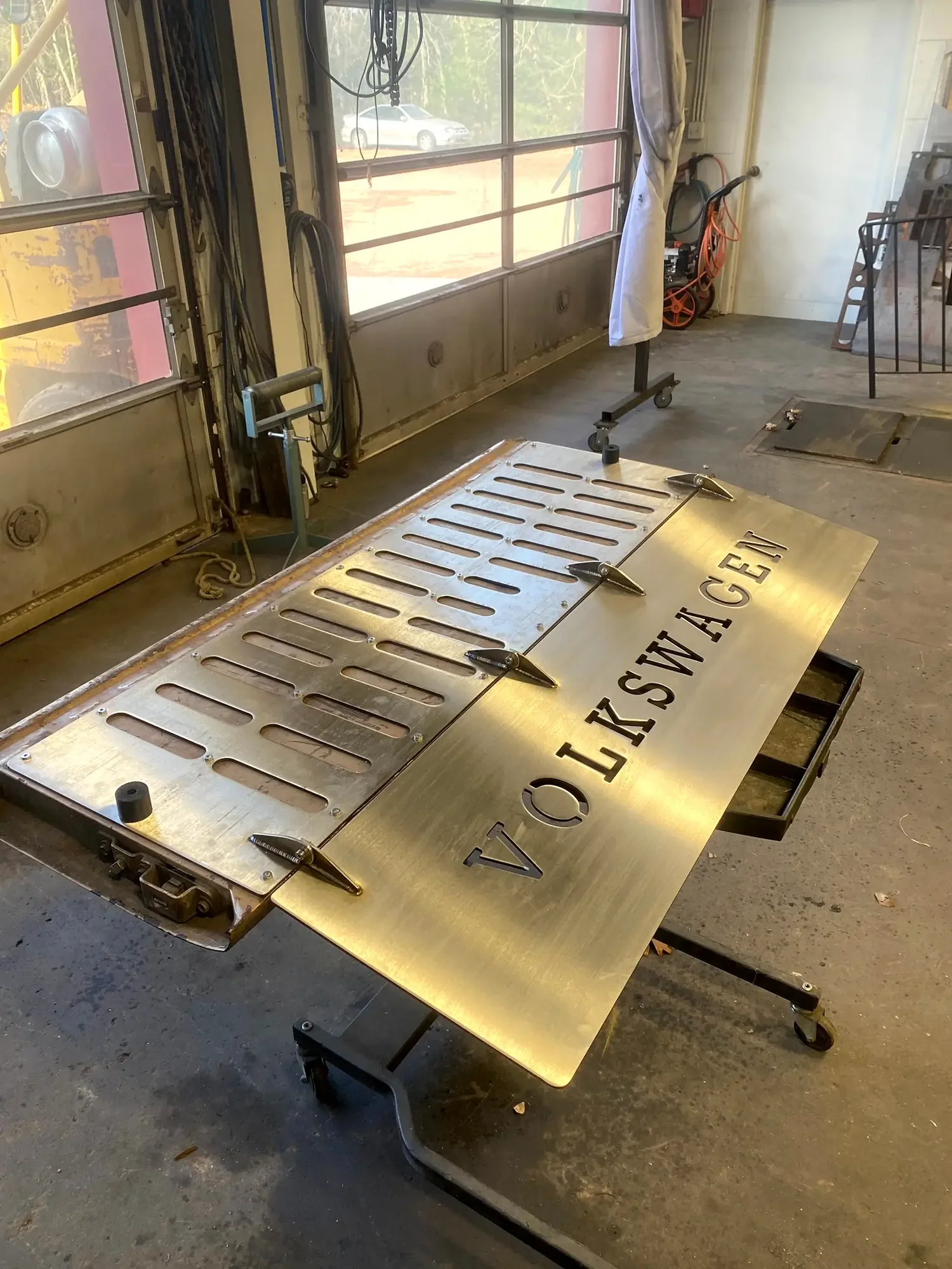 A large metal sign with the word 'ARKANSAS' cut out rests on a worktable in a workshop. The sign has slots for lights or letters and is secured with clamps. The workshop has a large window, tools, and equipment visible in the background.