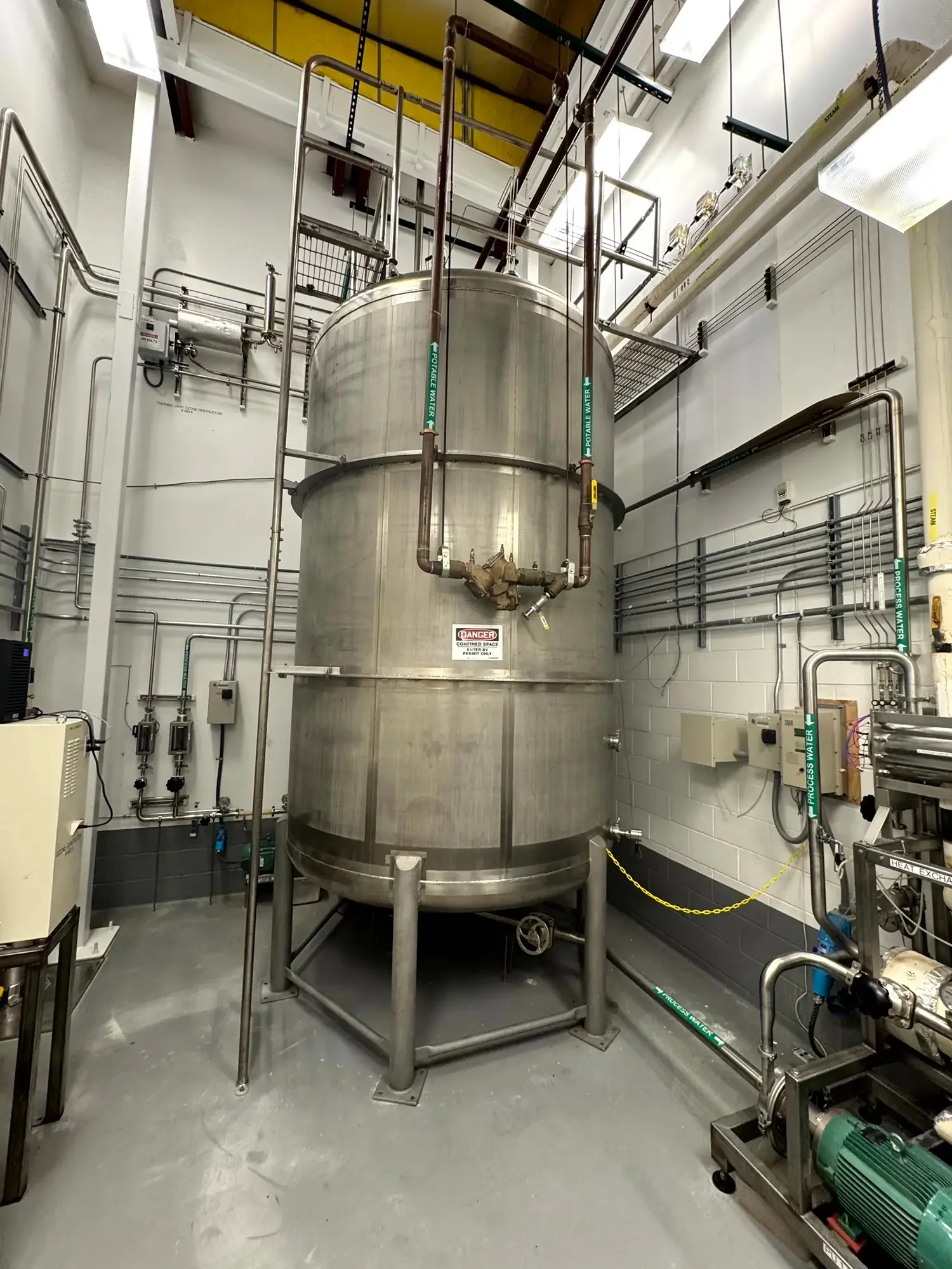 Large stainless steel tank in an industrial setting with pipes and gauges, surrounded by various equipment including a motor and control boxes.
