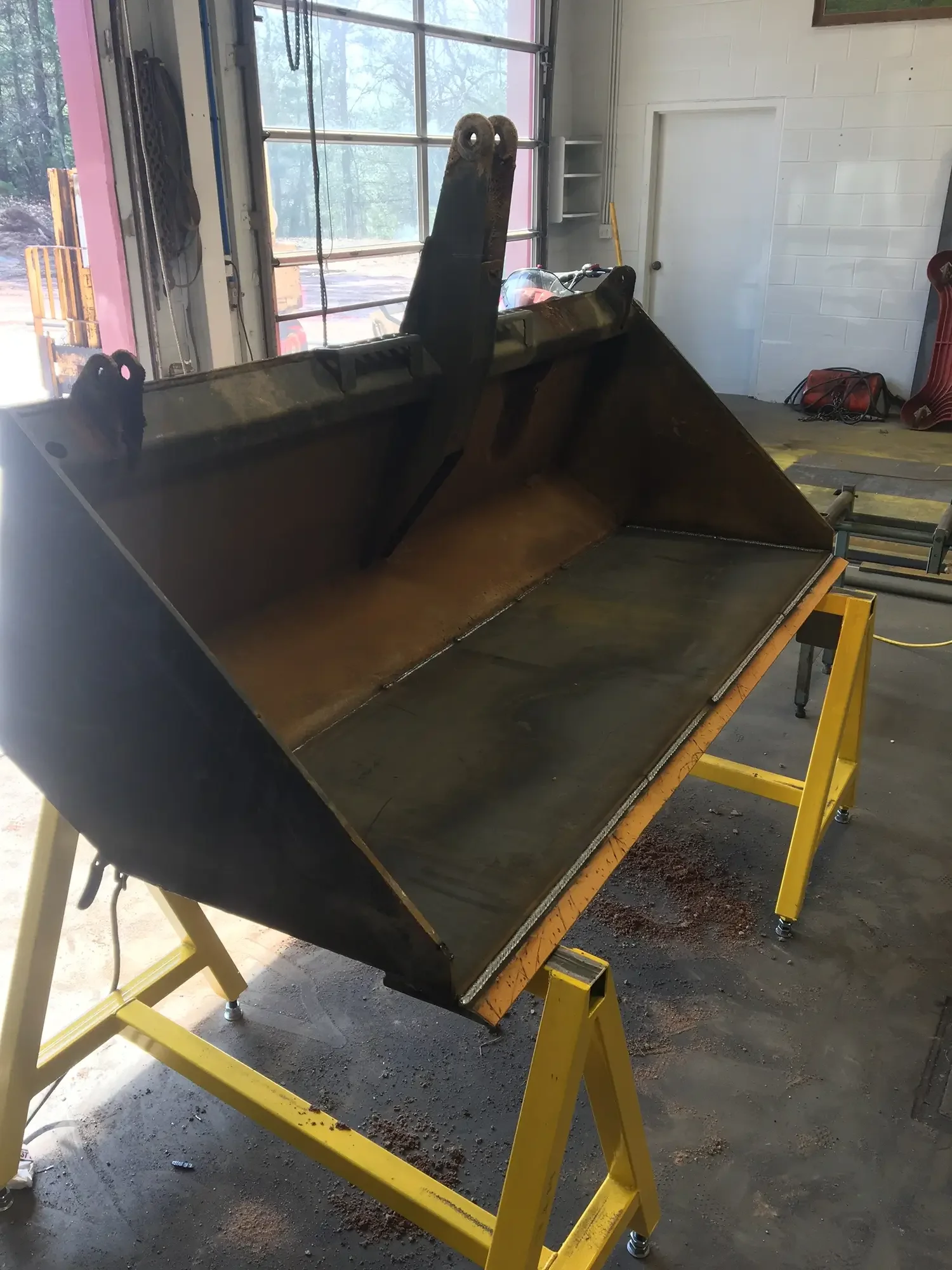 A large, black metal excavator bucket attached to a yellow stand in a garage or workshop, with a window showing trees outside and various tools and equipment inside.