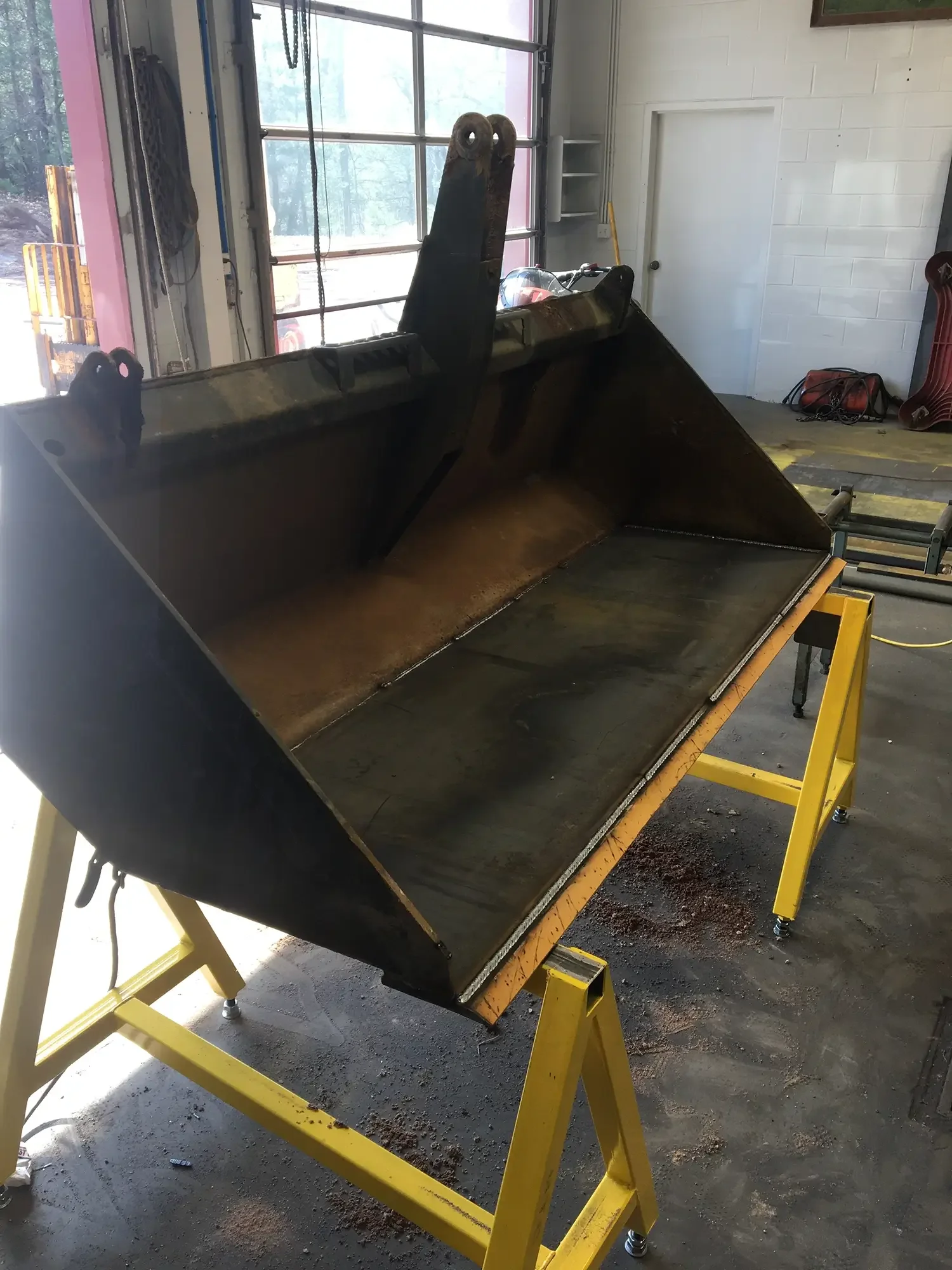 A large excavator bucket mounted on a yellow work table inside a workspace, with a garage door and a white brick wall in the background.