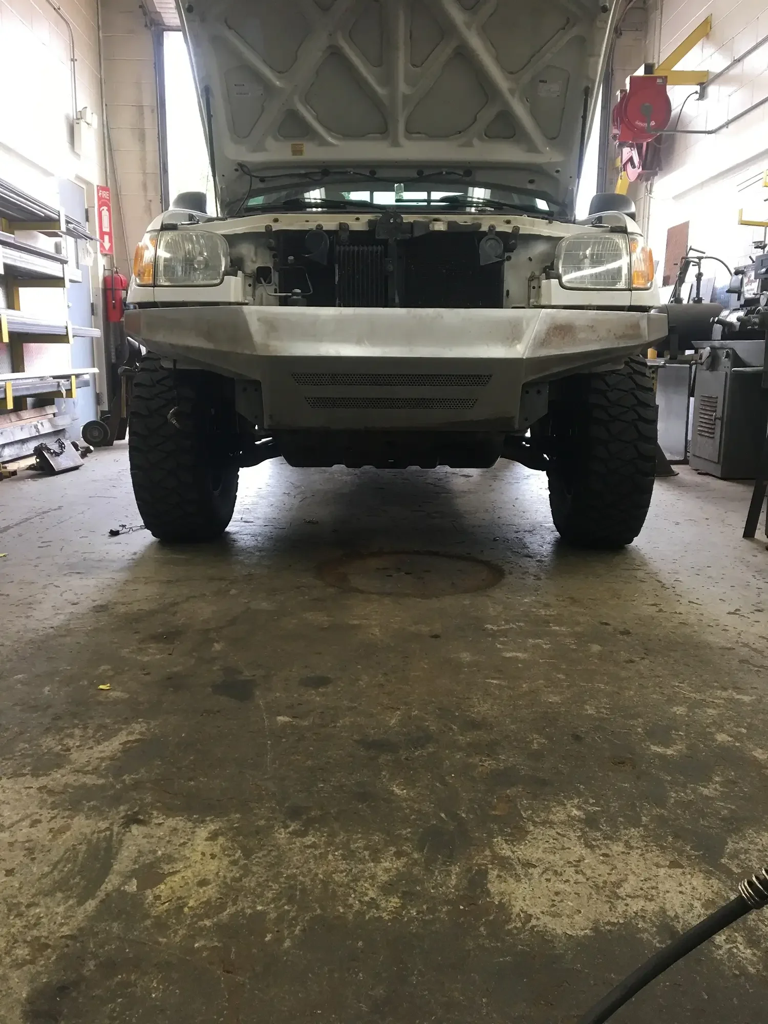 Front view of a white vehicle in a garage with the hood open, exposing the engine compartment. The garage has shelves, tools, and equipment along the walls.