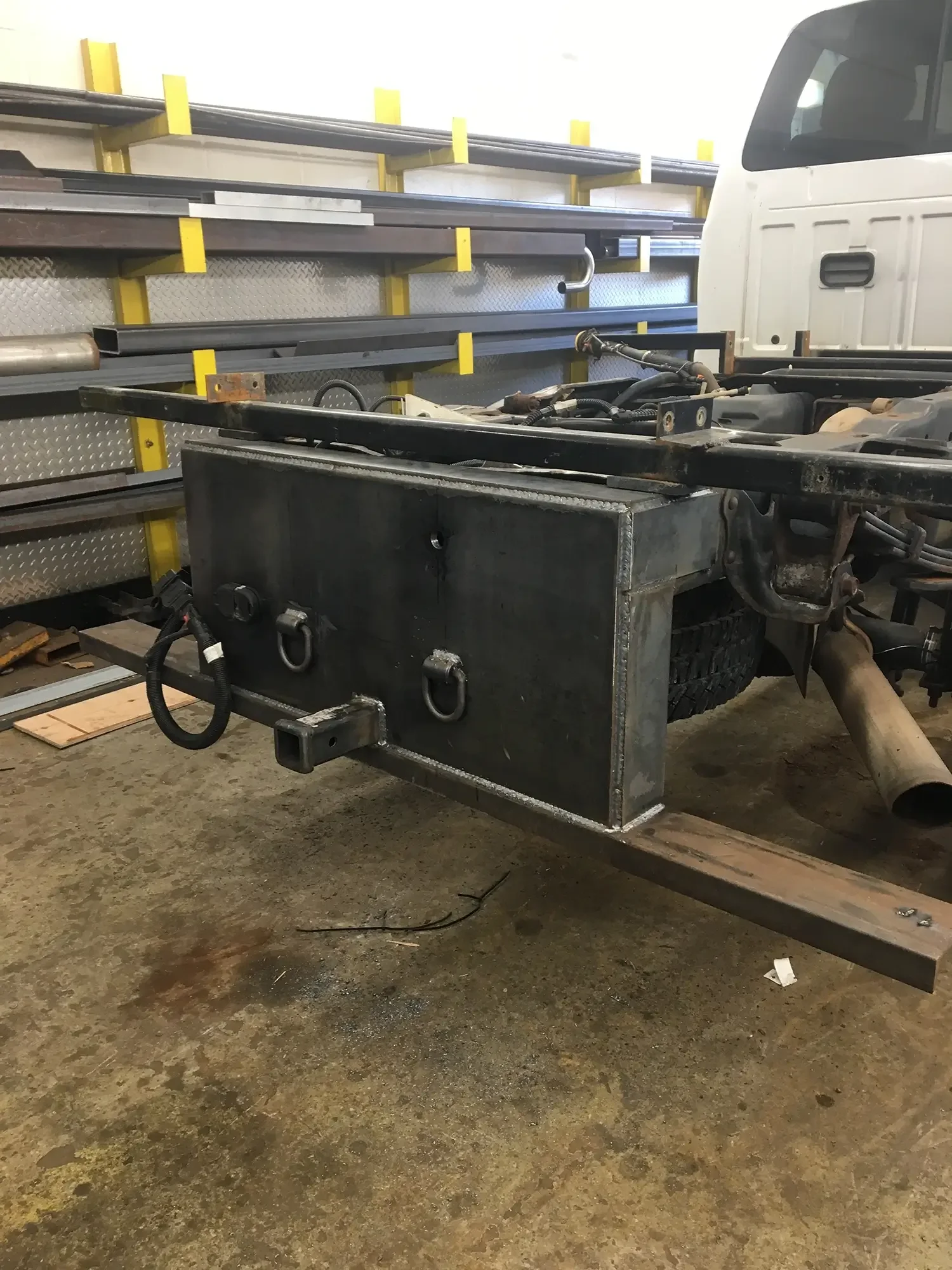 The image shows a partially assembled flatbed truck with a large metal platform, some tools, and parts on top, in a workshop with yellow and gray shelving in the background.