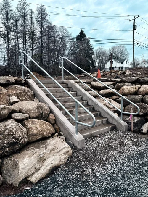 Concrete stairs with metal handrails leading up a small slope, surrounded by large rocks and gravel, with a background of leafless trees, utility poles, a house, and an orange traffic cone.