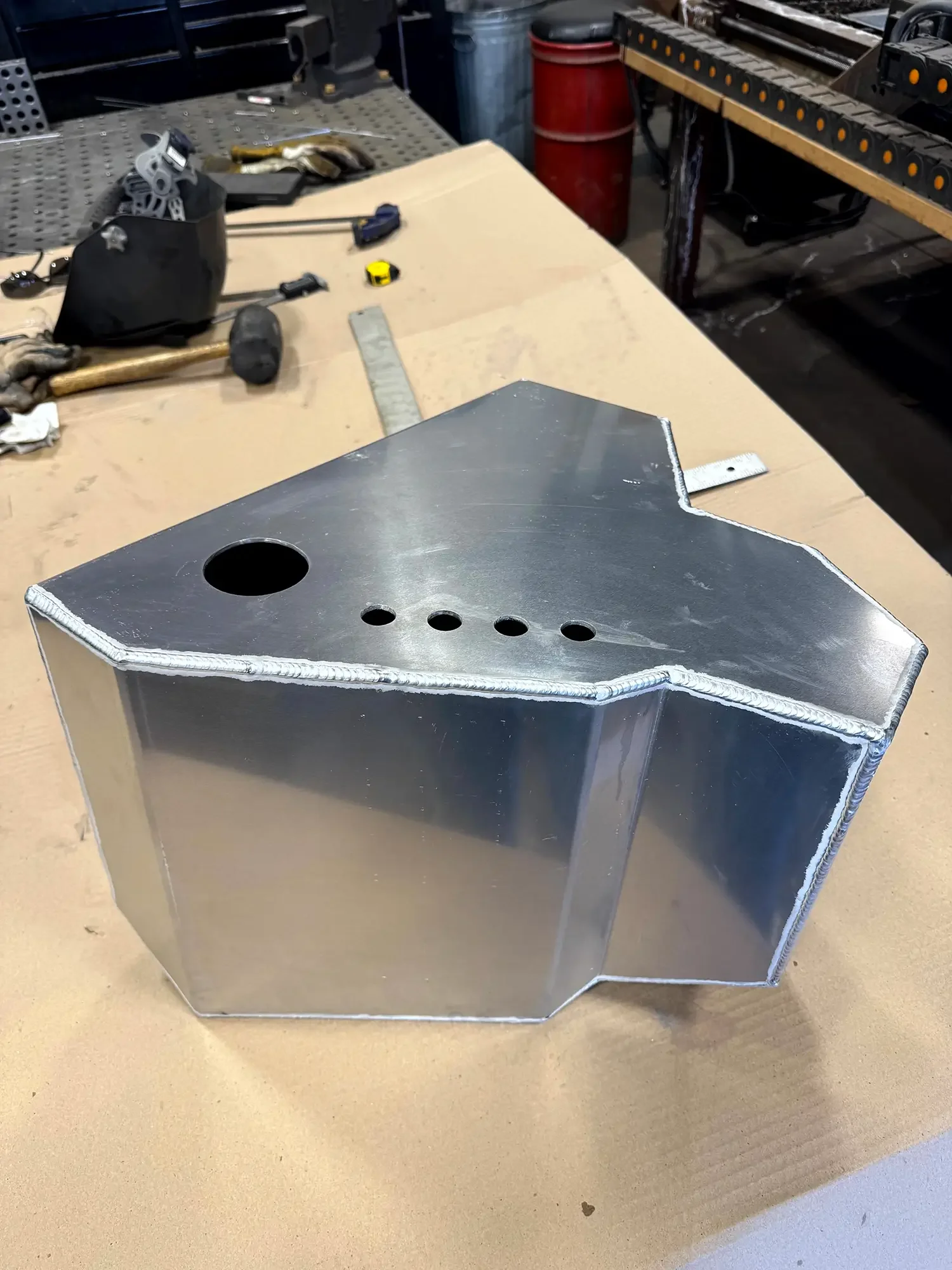 A metal welded box with holes on top, placed on a wooden workbench in a workshop. In the background, tools and equipment are visible.