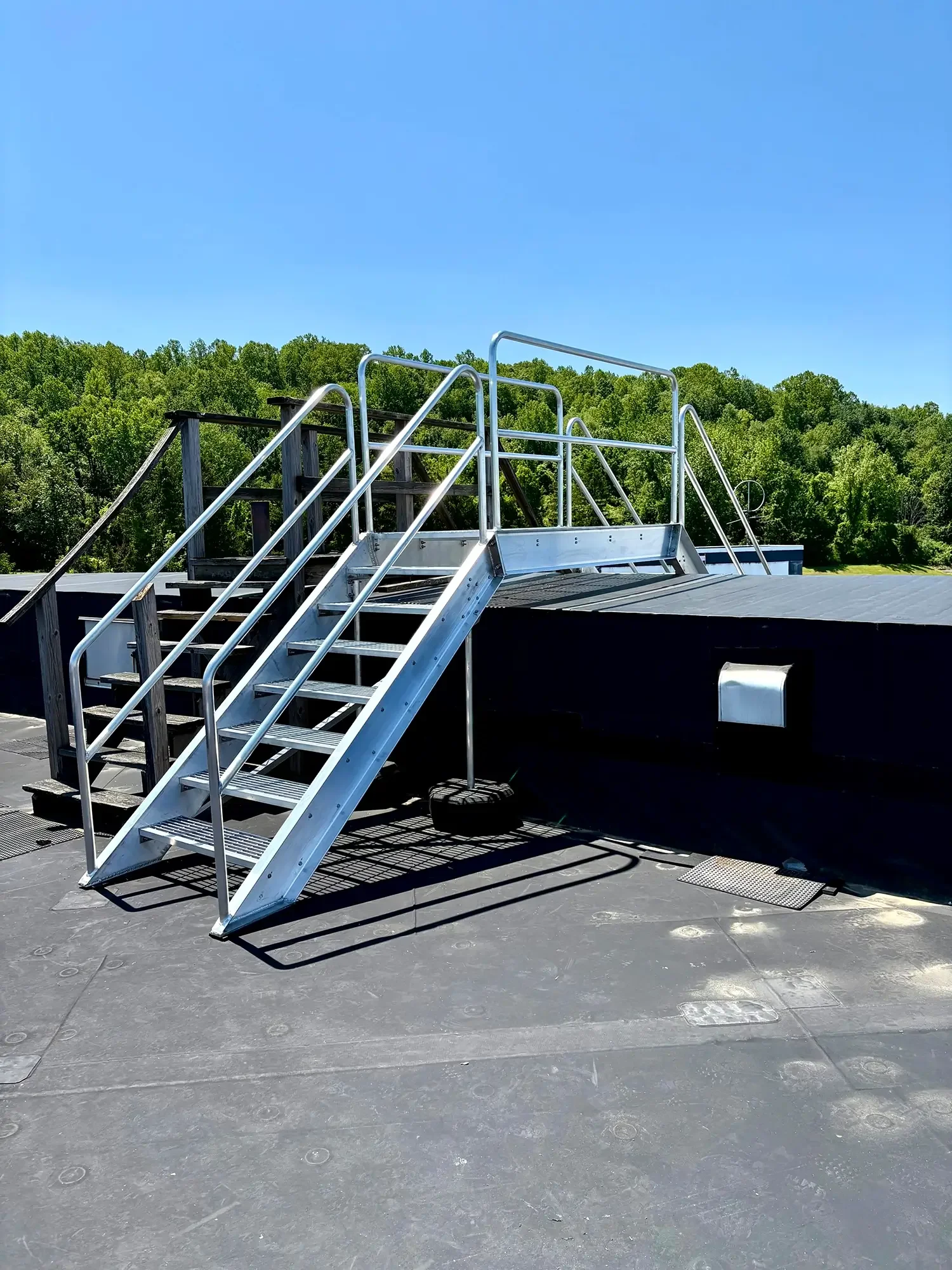 Metal stairs leading to an outdoor platform on a rooftop with a railing, under a clear blue sky with trees in the background.