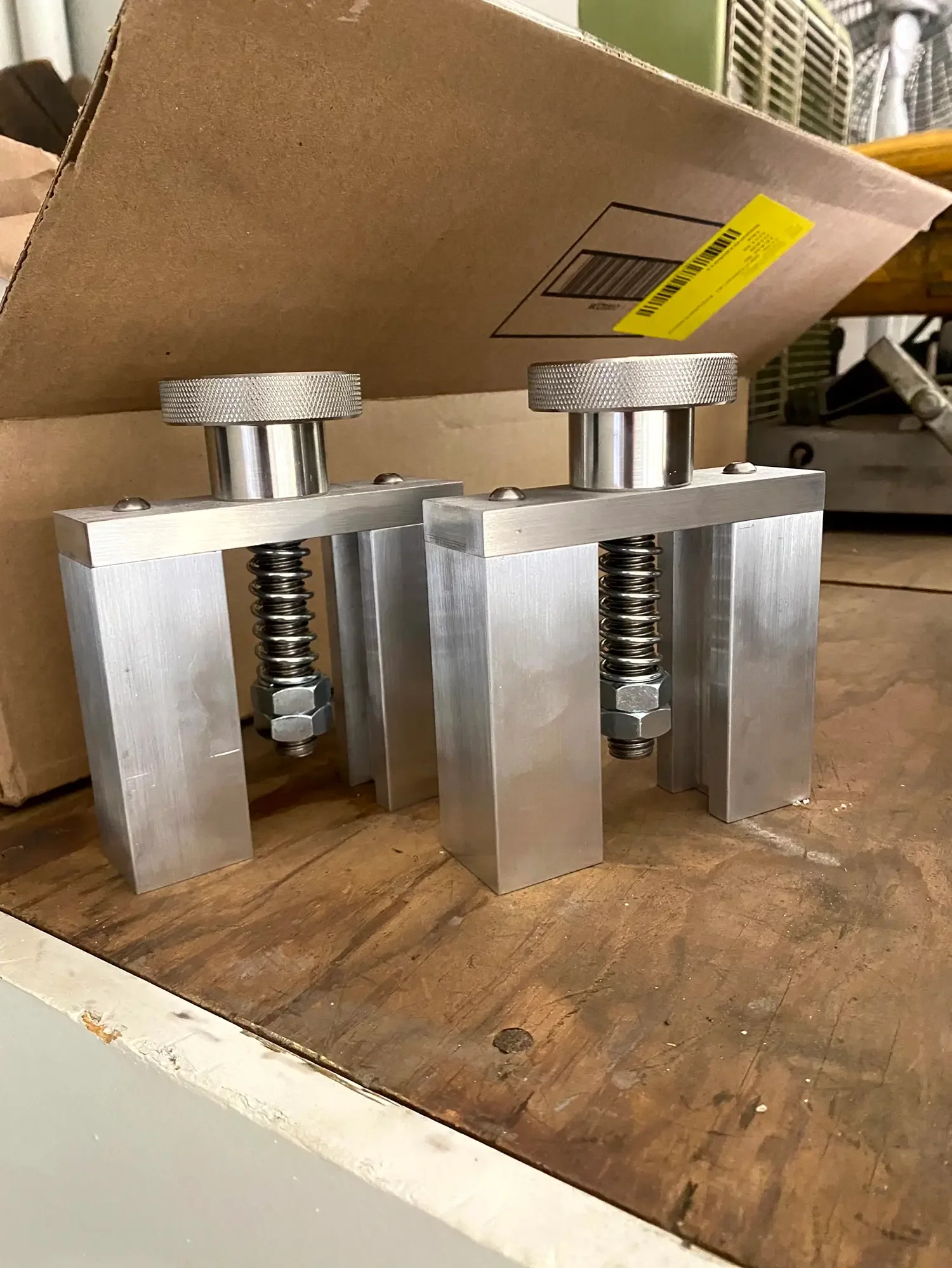 Metal device with two vertical adjustable components and springs, placed on a wooden surface with a cardboard box in the background.