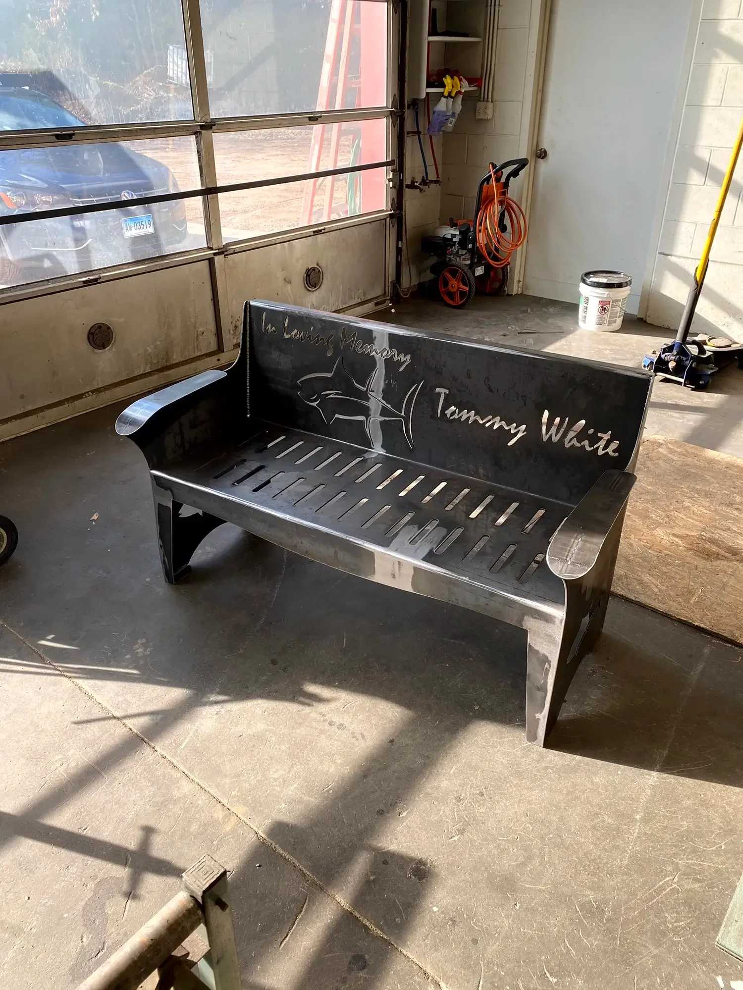 A black metal bench with the words "In Loving Memory Tommy White" written on the backrest, placed on a concrete floor in a garage. The garage has a large window and various tools, including a portable air compressor and a container of paint.