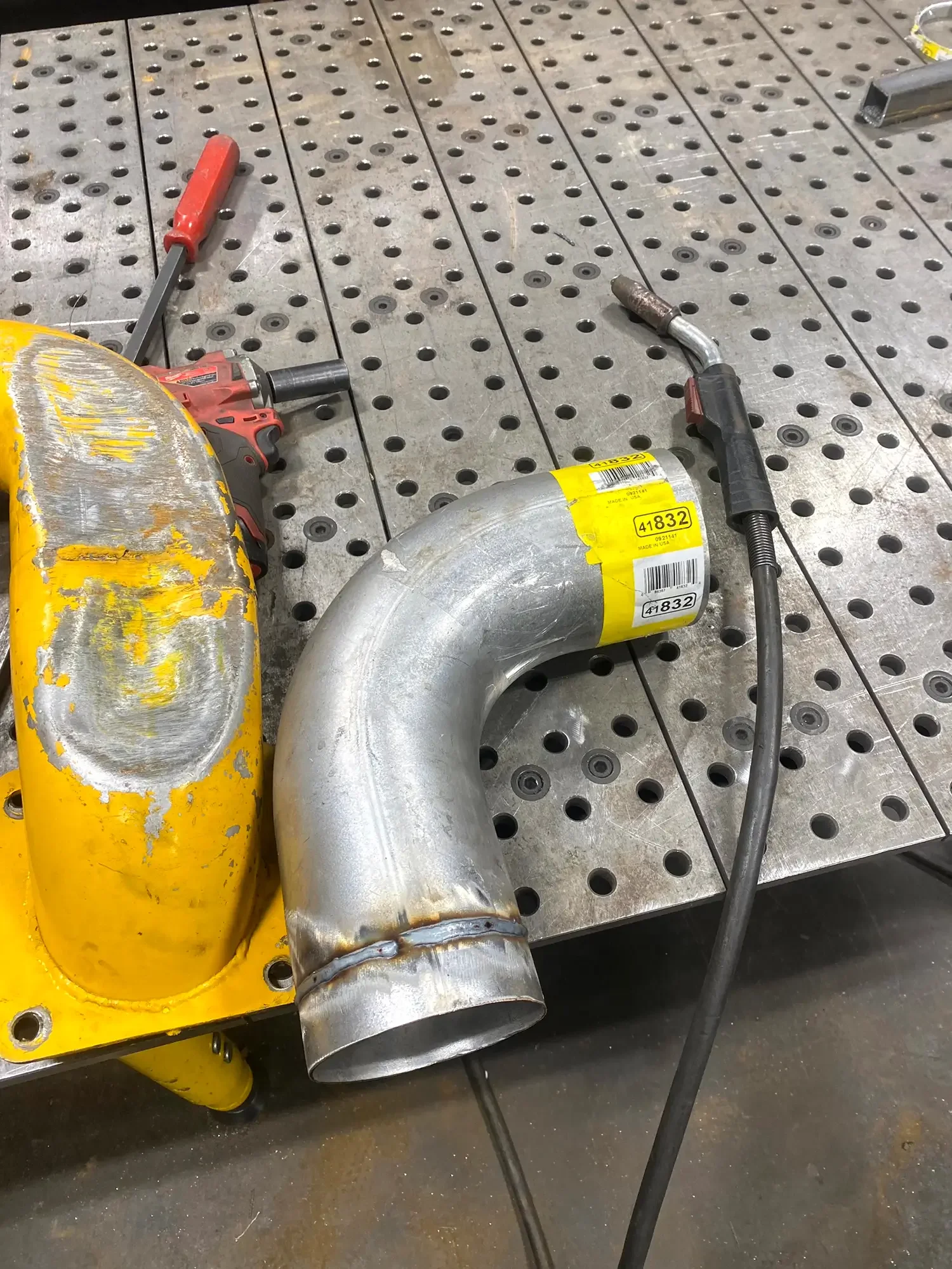 Metal pipes and tools on a perforated metal workbench in a workshop. The pipes are curved, with one showing yellow rust paint and another with a yellow barcode label.