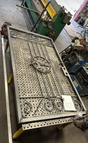 A large, ornate metal bed frame with decorative scrollwork is placed on a table in a workshop. The frame features a circular design in the center and intricate patterns on the head and foot sections.