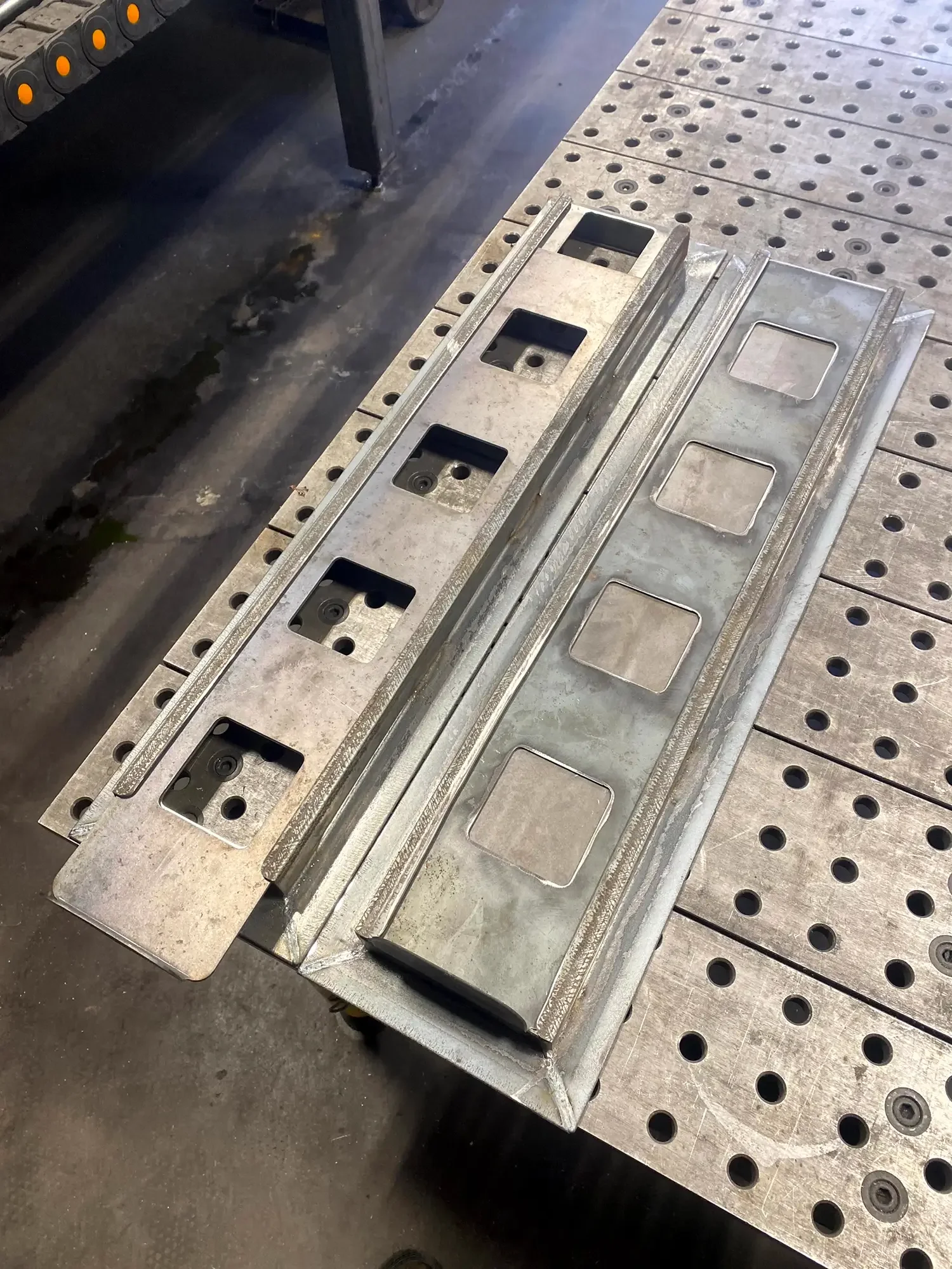 Metalworking workbench with tools and metal parts, including a long metal piece with square slots and holes, on a perforated metal table in an industrial workshop.