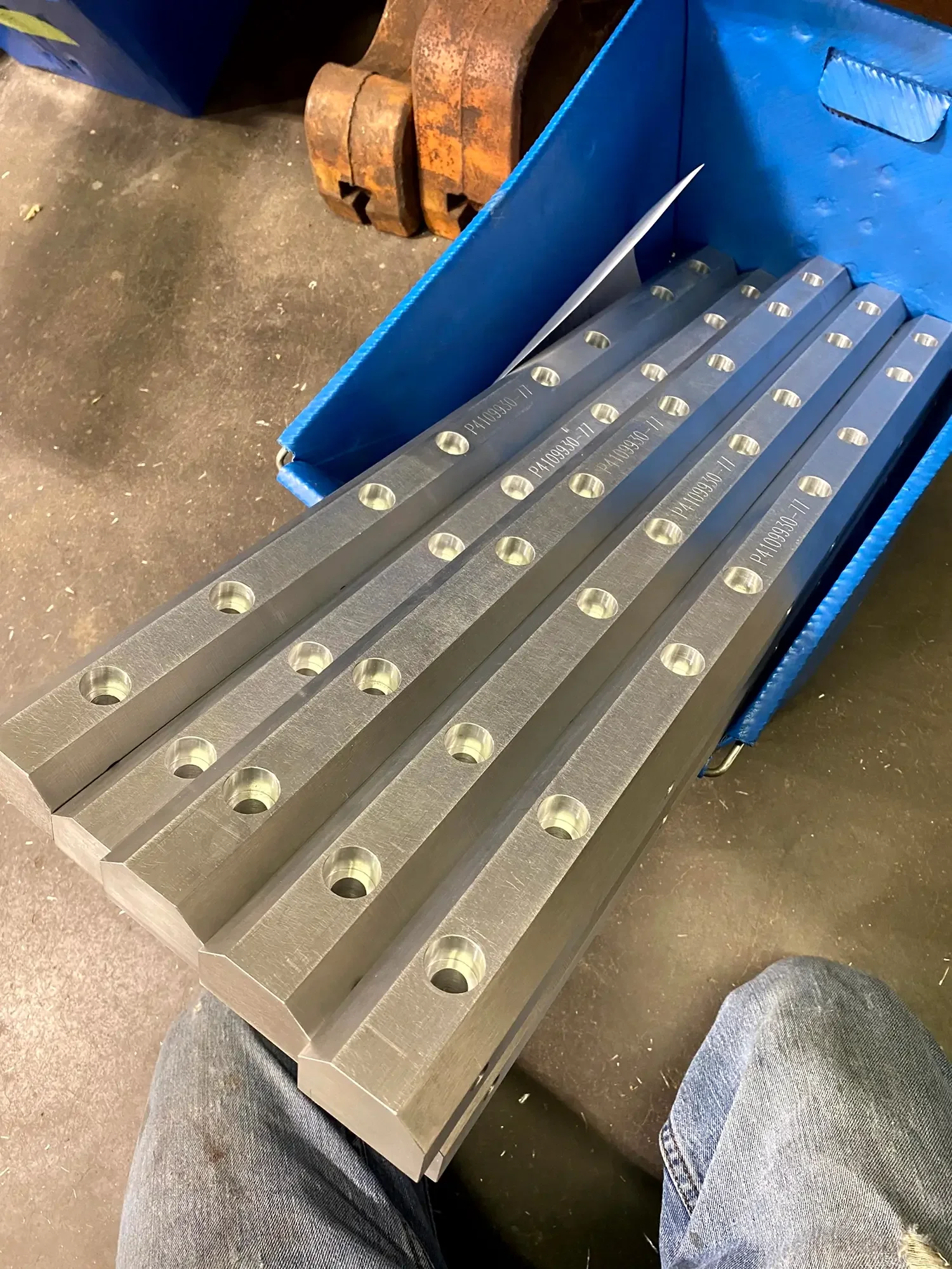A blue metal storage bin filled with long silver metal pieces with multiple holes, labeled with the code P4109930-11. The bin is on the floor, with parts of a person's legs and jeans visible at the bottom.