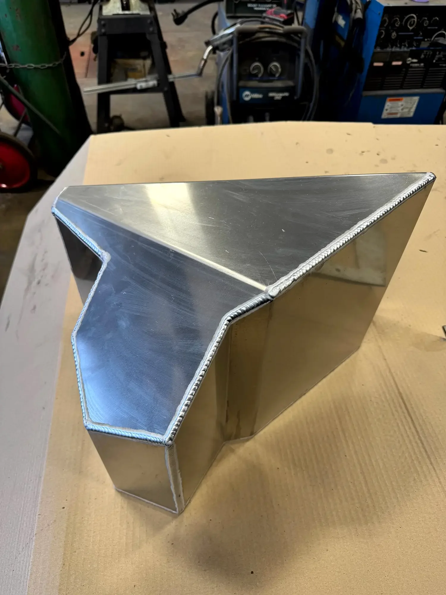 A metal, angular, L-shaped object with a welded seam along the edges sitting on a wooden workbench in a workshop.