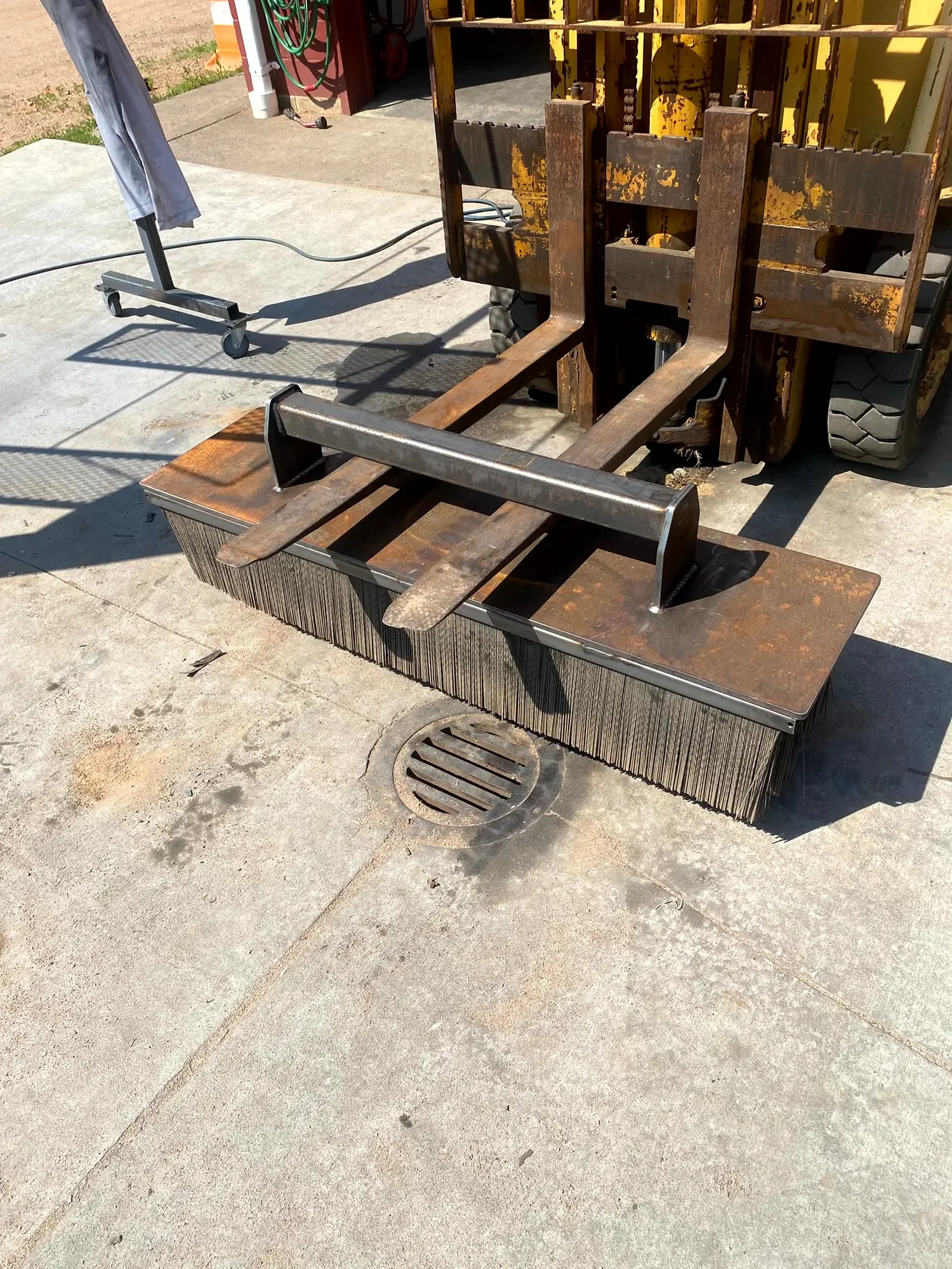 A forklift with rusty forks parked outside on concrete ground near a drain, with a clothes rack and some garden hoses in the background.