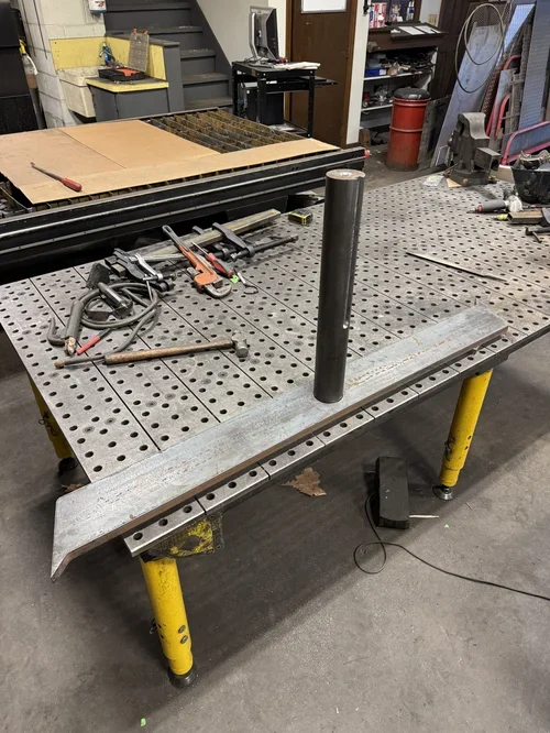 A workshop workspace with a large metal work table with yellow legs, holding various tools including clamps, a hammer, and a square. A black pipe stands upright on the table. In the background, there are shelves, a computer, and other workshop suppli