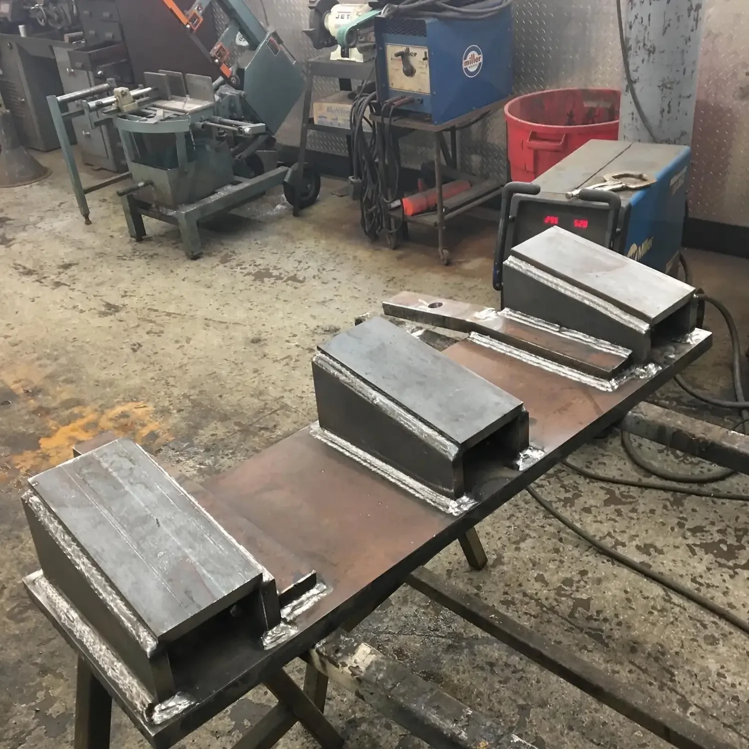 Metalwork workspace with welded aluminum pieces on workbenches, welding machine, tools, and equipment in an industrial setting.