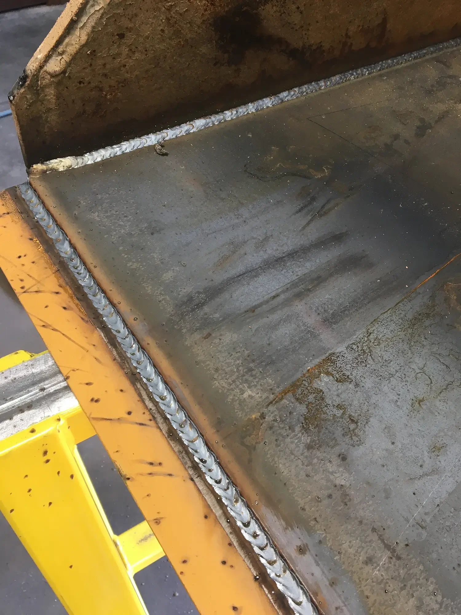 Close-up of a welded metal corner joint on a large industrial metal surface, with orange safety barrier in the foreground.