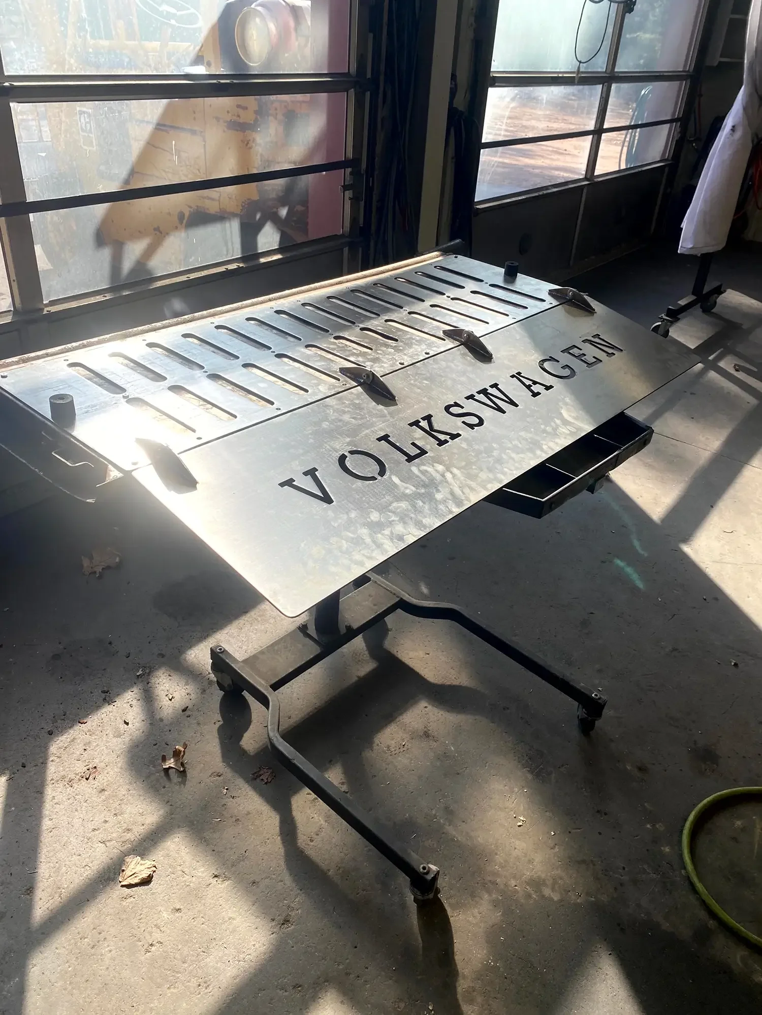 A metal stretcher with the word 'VANKSWAGEN' engraved on it, placed on a concrete floor with some scattered leaves, near a window with sunlight streaming in.