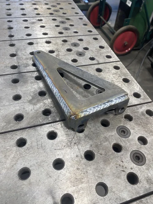 A metal welding fixture with a cutout handle, placed on a perforated workbench, showing a completed welded seam around the edge.