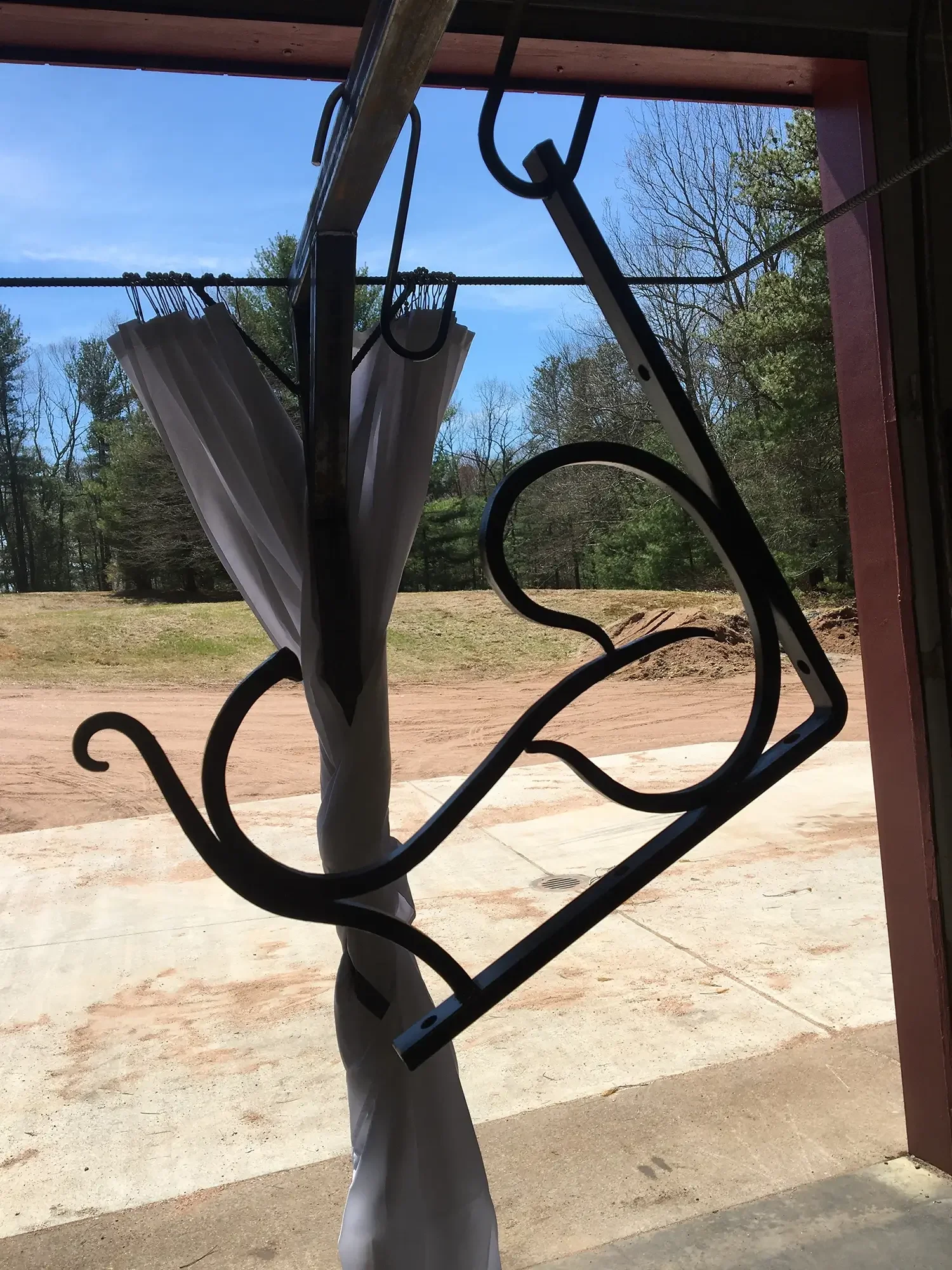 View through a decorative black wrought iron gate, showing a patio with a concrete surface, a dirt yard, trees, and a clear blue sky.