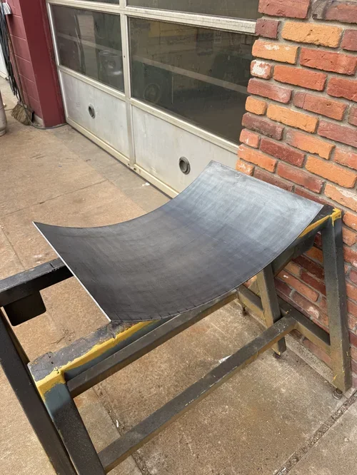 A curved, black skateboard ramp mounted on a wooden stand outside a building with a brick wall and garage door.