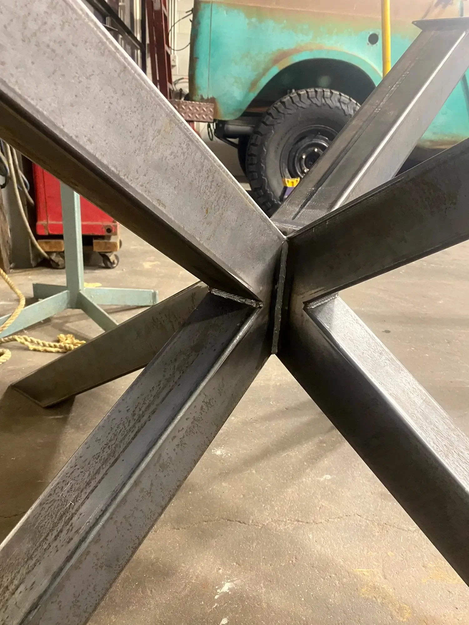 Close-up of a steel cross structure with multiple beams meeting at a central point, with a work area and tools in the background.